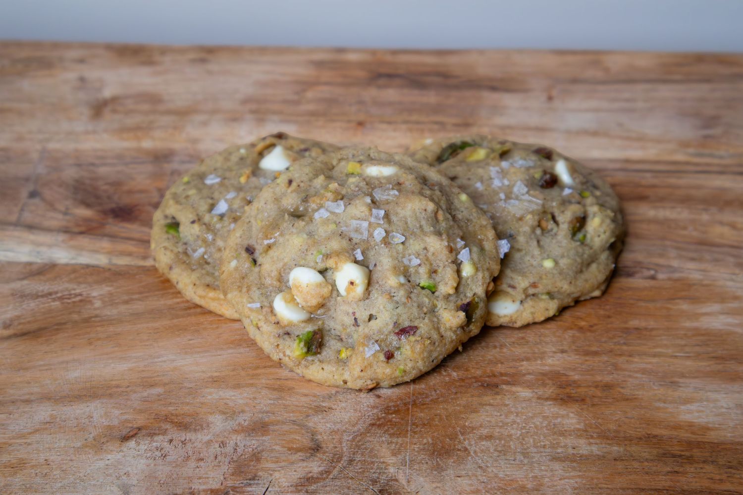 Three pistachio white chocolate chip cookies with sea salt on a wooden cutting board.