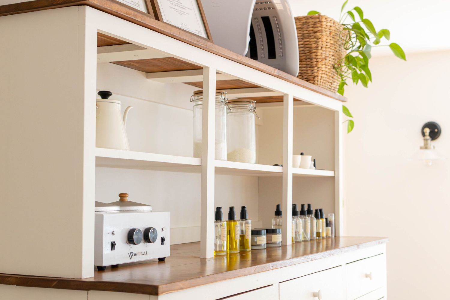 White shelving unit displaying glass bottles, oils, and wax warmer in a bright spa or salon treatment room.
