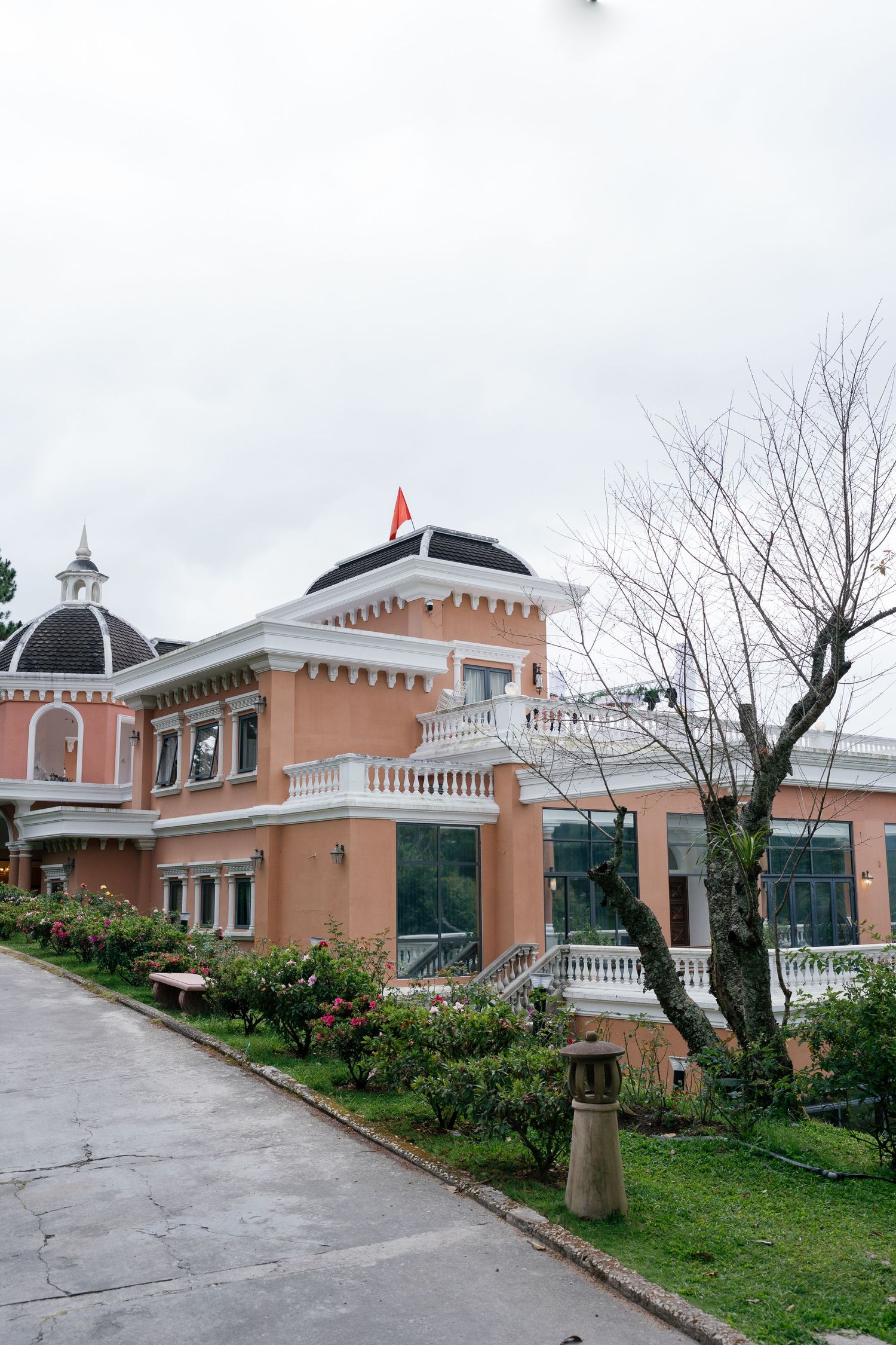 Pink colonial-style building with white balconies, dome roof, and Vietnamese flag, surrounded by manicured gardens.