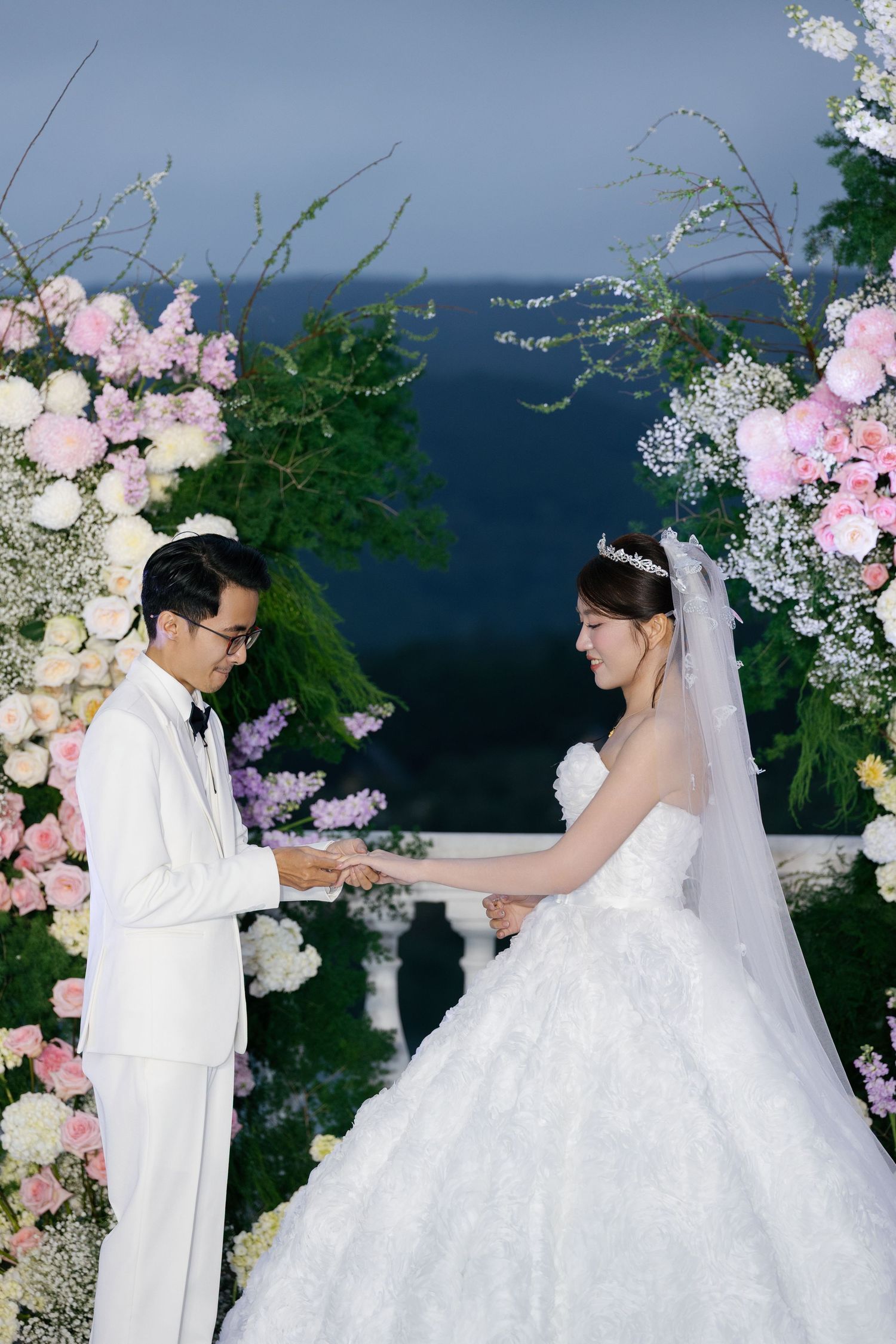 Couple exchanging rings at an outdoor wedding ceremony surrounded by pink and white floral arch with lush greenery.