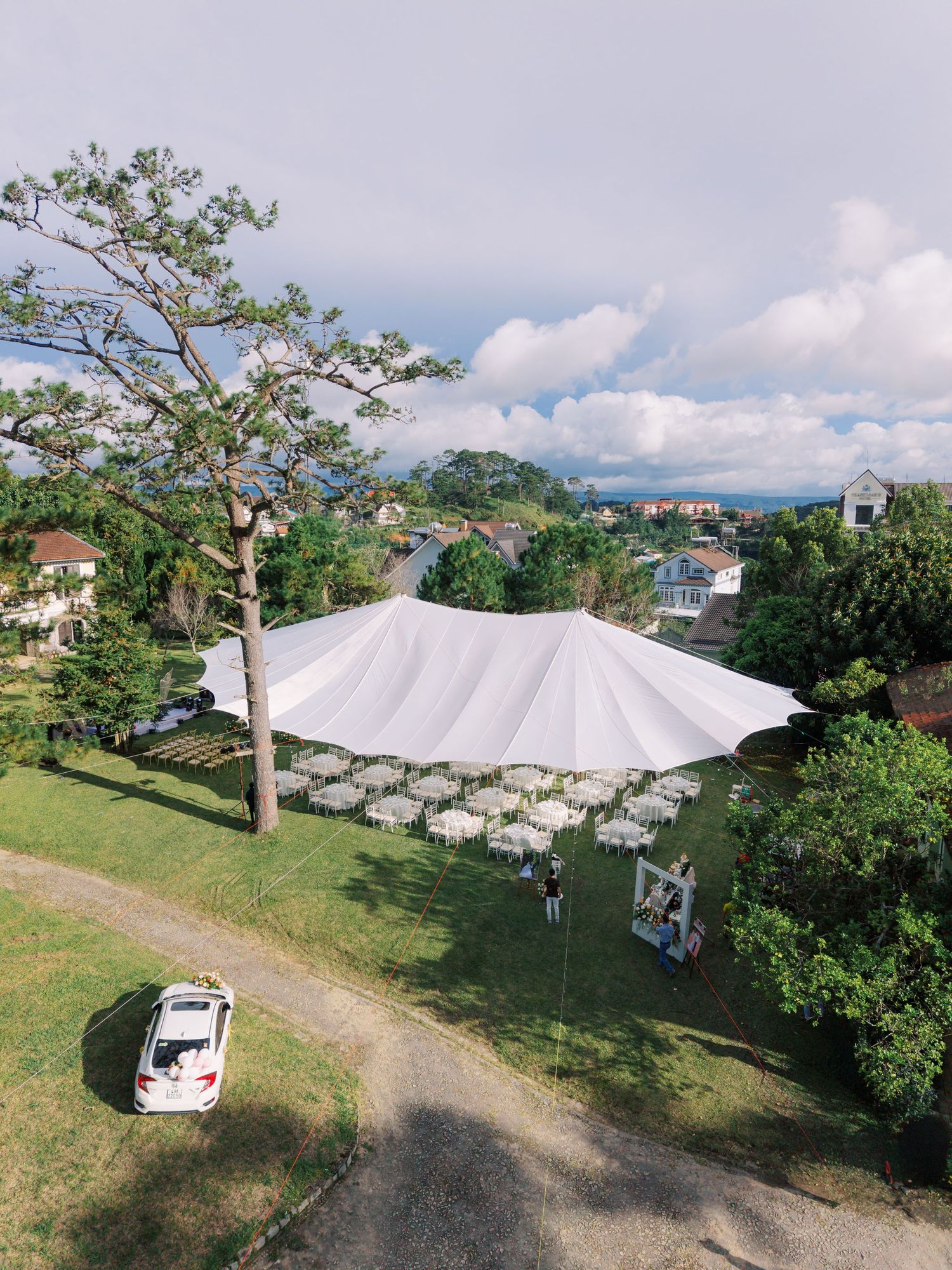 Aerial view of a large white event tent set up on a green lawn surrounded by trees and residential buildings under a cloudy sky.