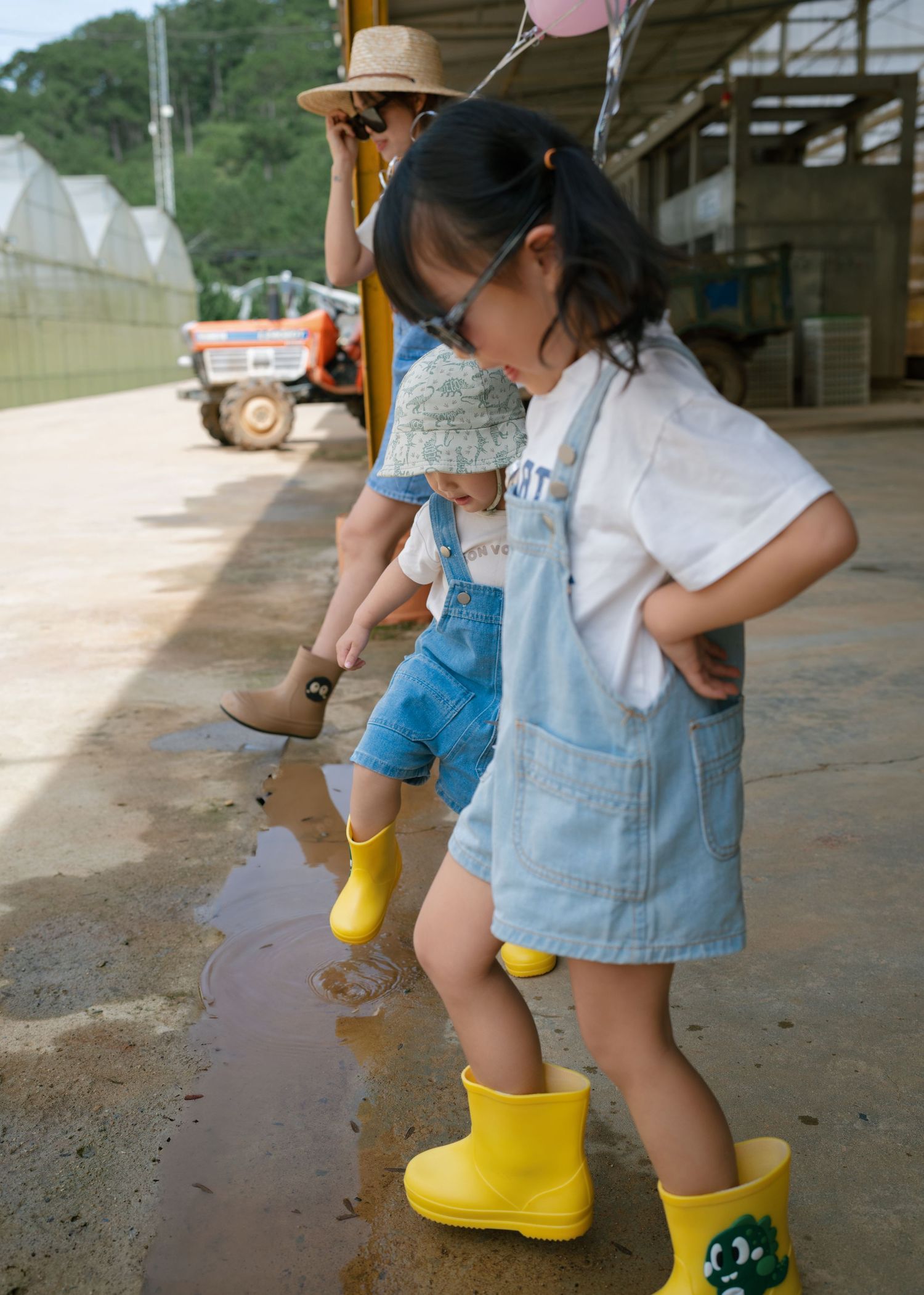 Two young children in yellow rain boots and denim overalls splash in puddles at a farm, with an adult nearby.