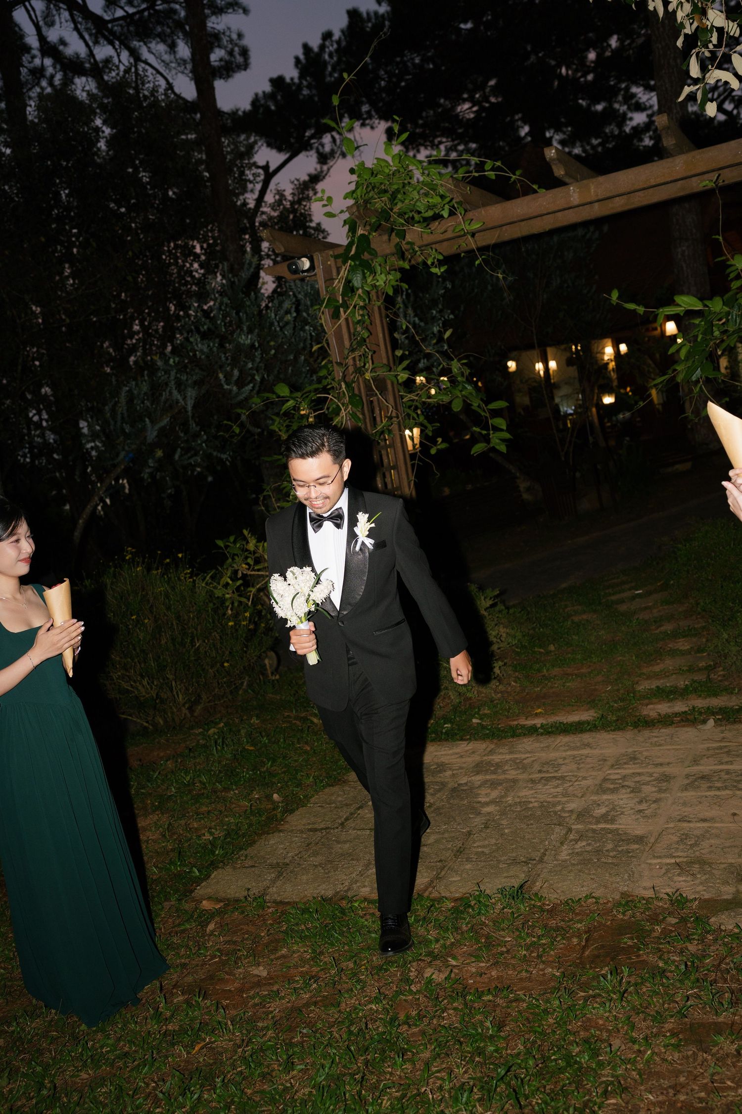 Groom in black tuxedo with bow tie carrying white bouquet walking down garden path at night.