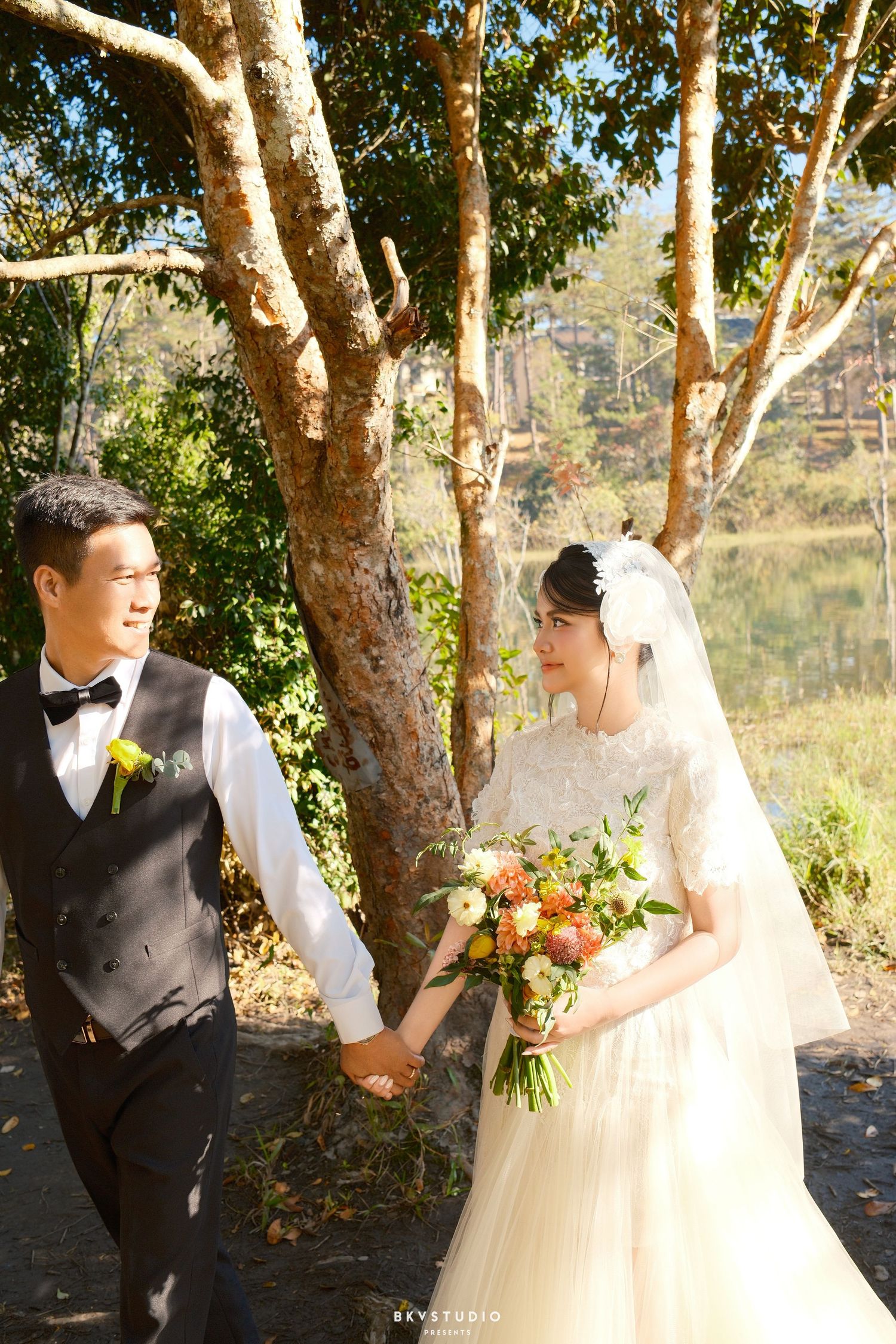 Smiling wedding couple holds hands near large tree, bride holding colorful bouquet in sunlit park.