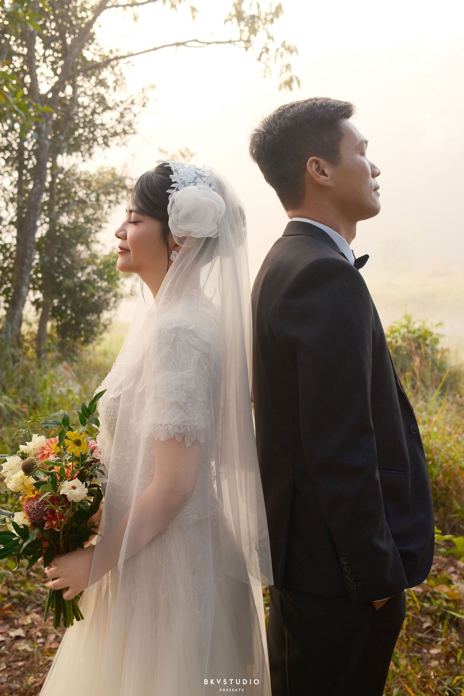 Bride and groom stand back-to-back in misty forest, bride holding colorful bouquet in white lace gown with veil.