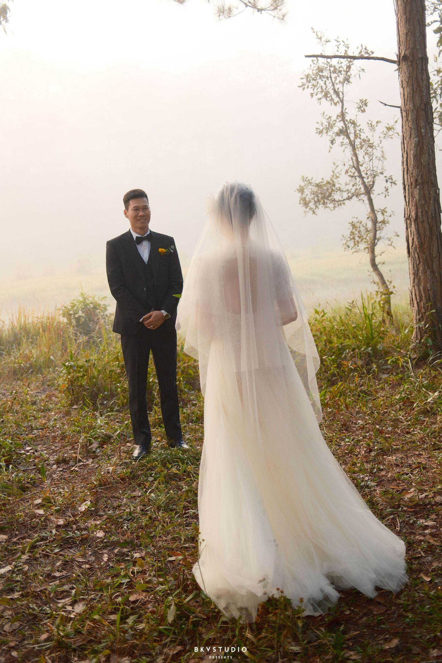 Groom in black tuxedo smiles at bride in white wedding gown and veil during misty outdoor first look.