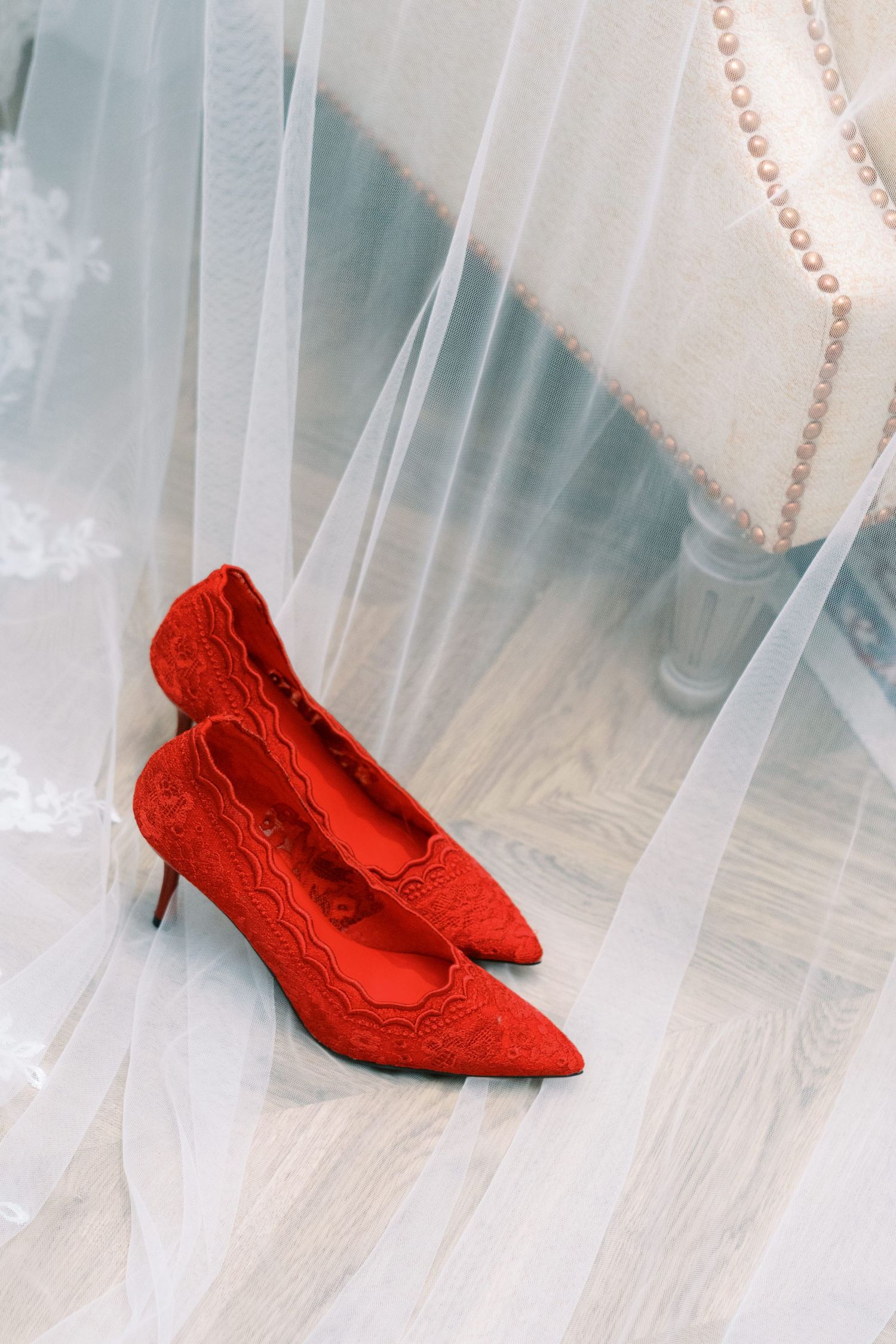 Red lace pointed-toe kitten heels placed on white tulle fabric with pearl necklace and upholstered chair in background.