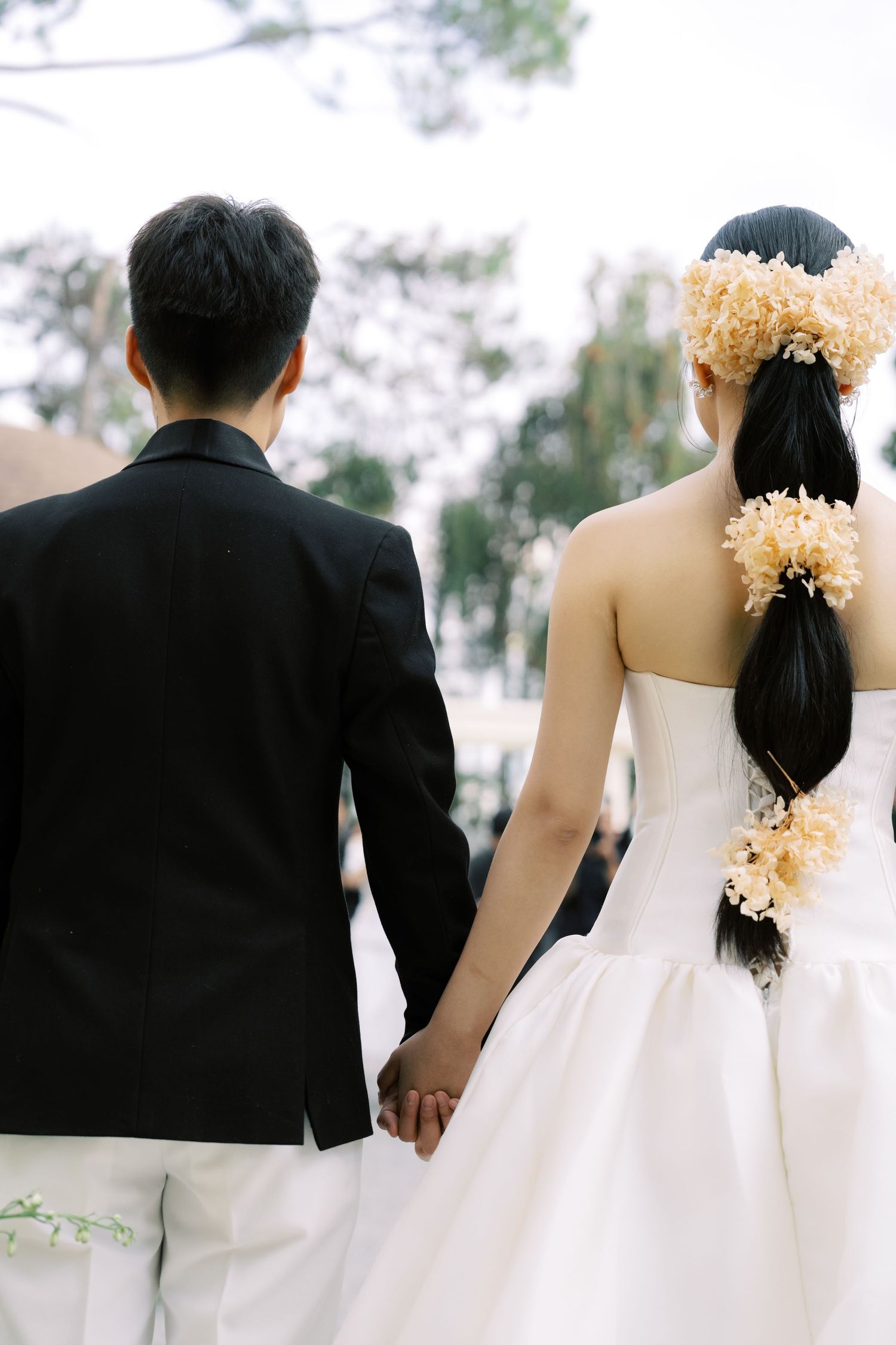 Bride and groom hold hands from behind, bride wearing floral hair accessories in her dark ponytail.
