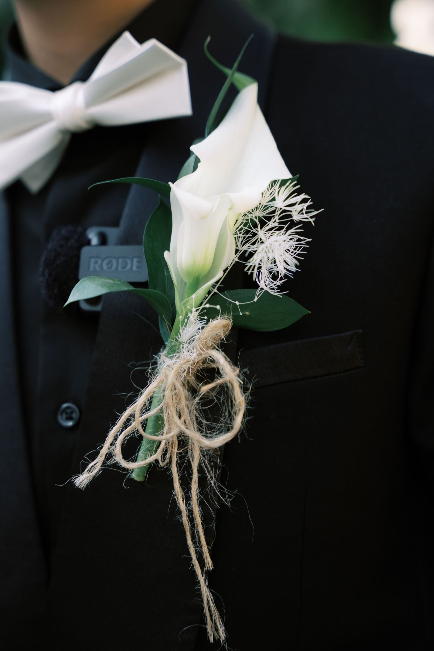 Groom's black tuxedo lapel with white calla lily and twine boutonniere and white bow tie.