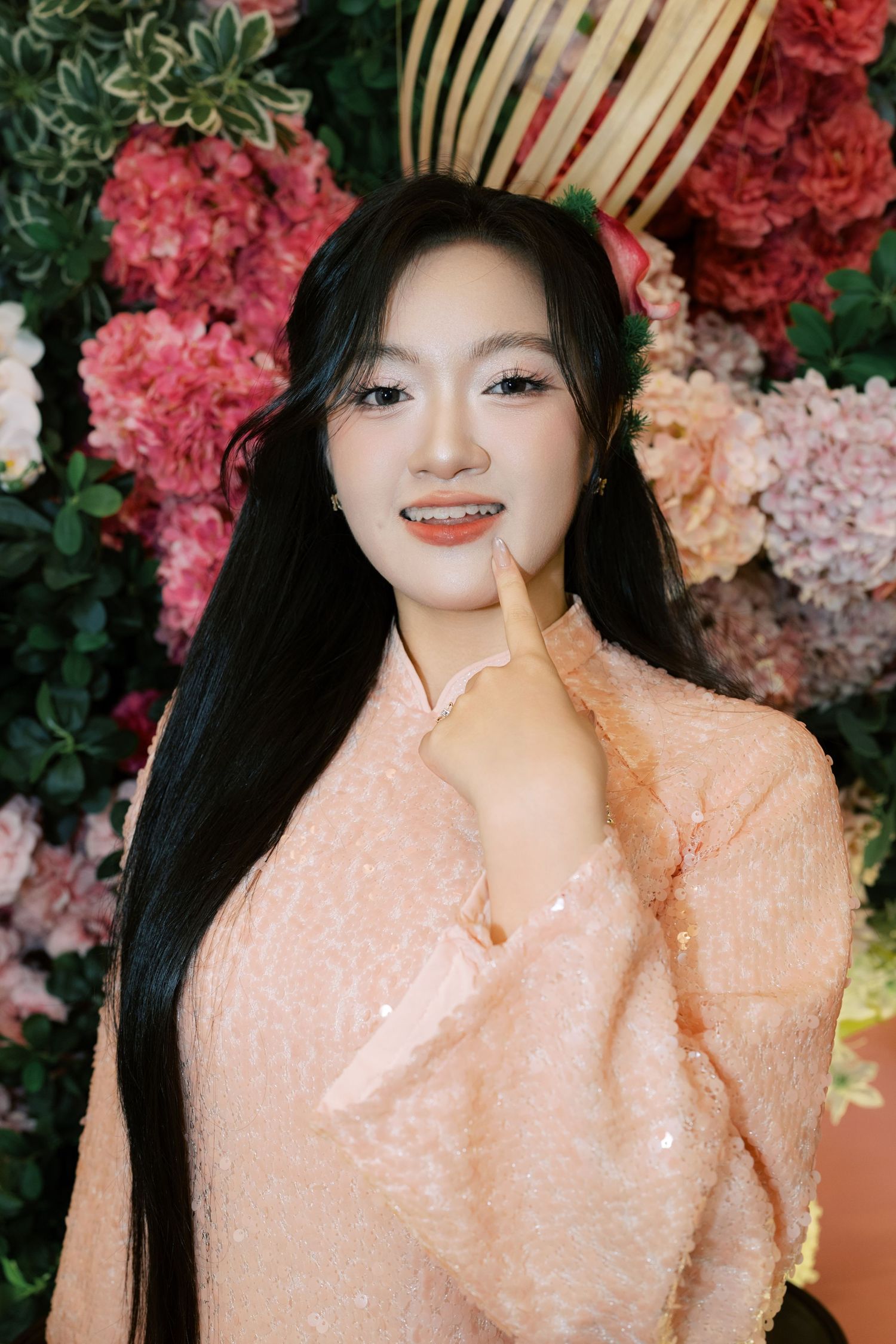 Young woman in pink sequin dress poses gracefully in front of a vibrant floral wall backdrop.