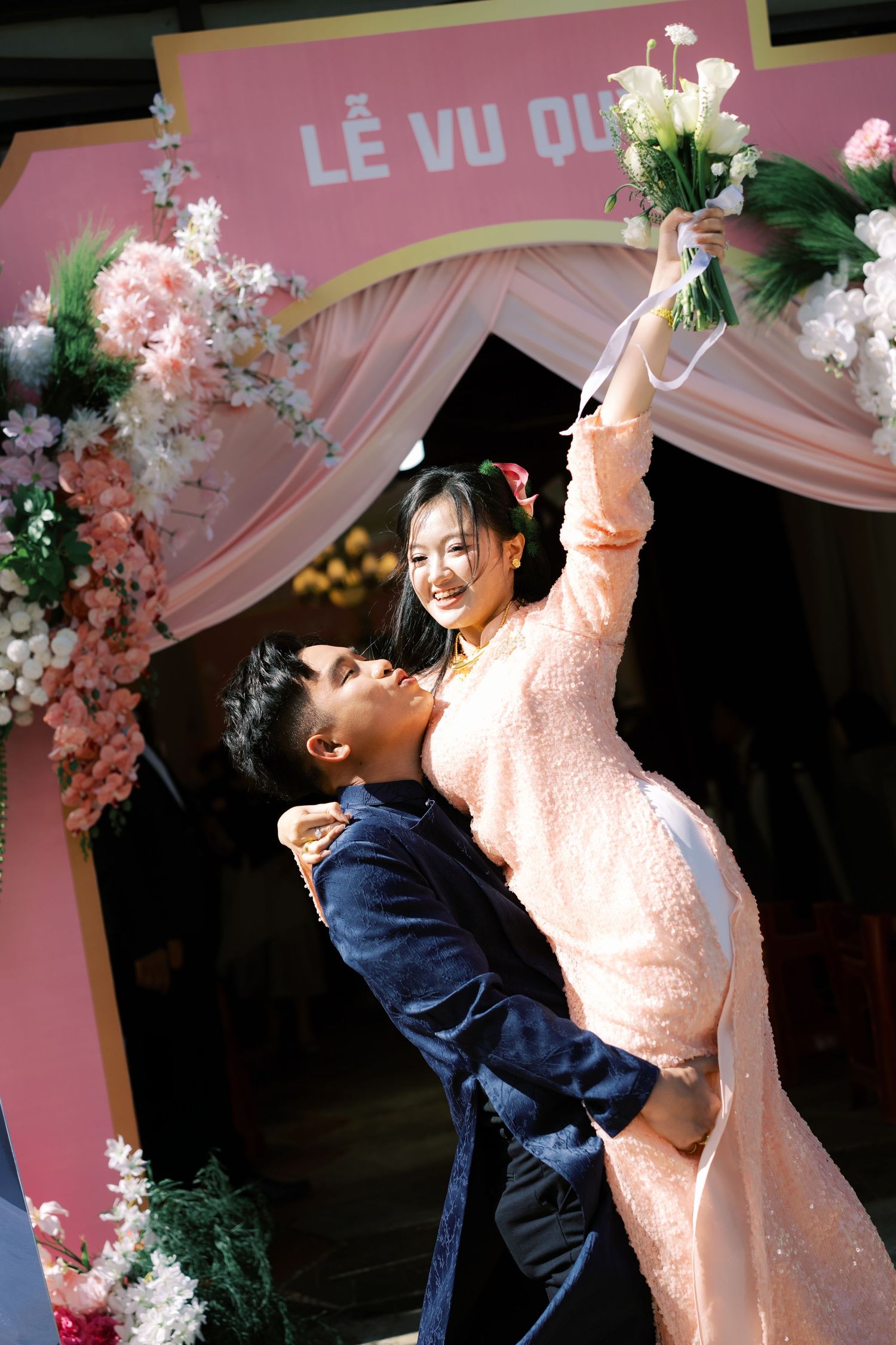 Joyful Vietnamese bride in pink ao dai lifted by groom under floral wedding arch at Le Vu Quy ceremony.