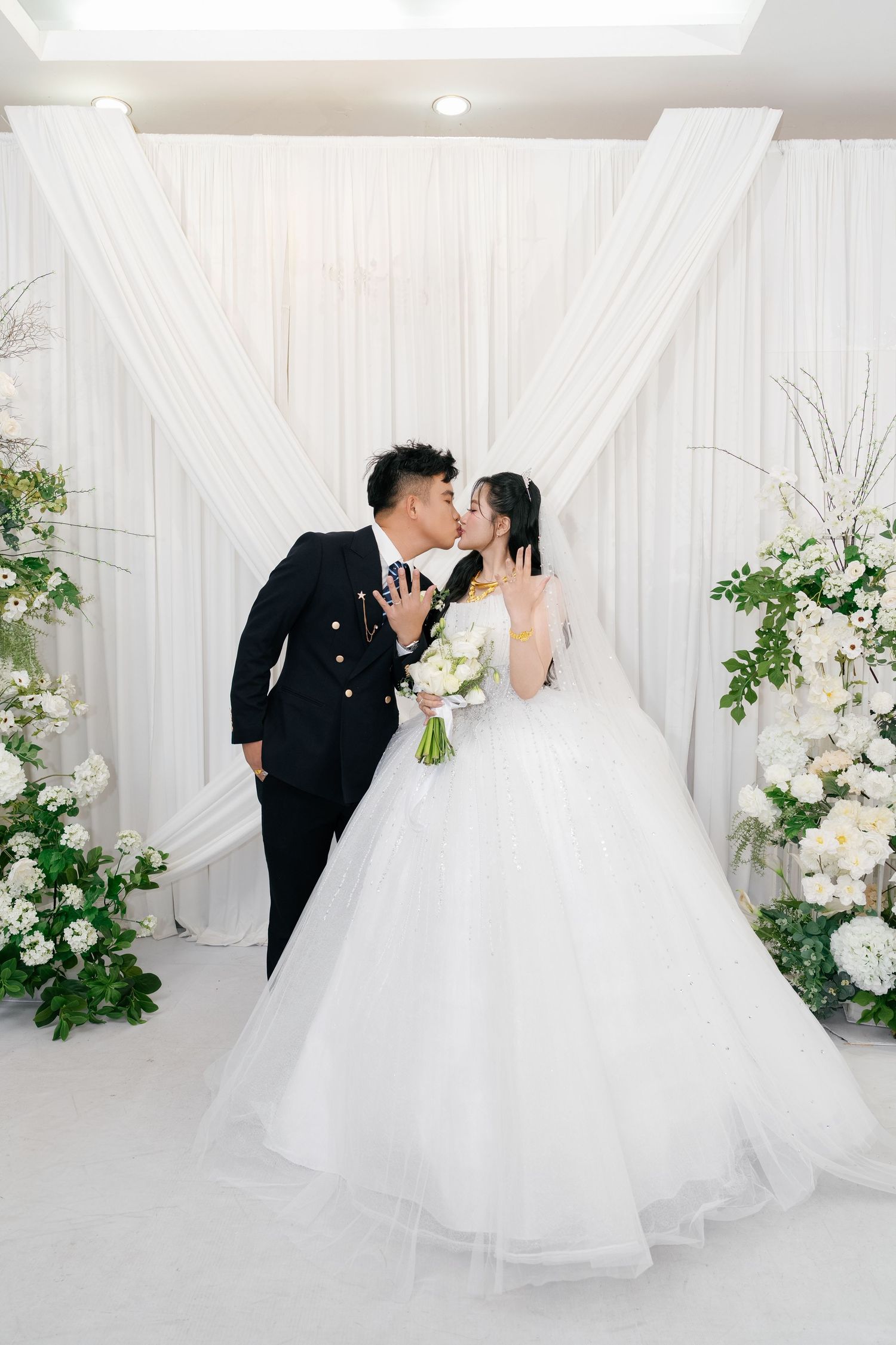 Bride and groom share a kiss at elegant white floral wedding altar, bride in ball gown holding bouquet.