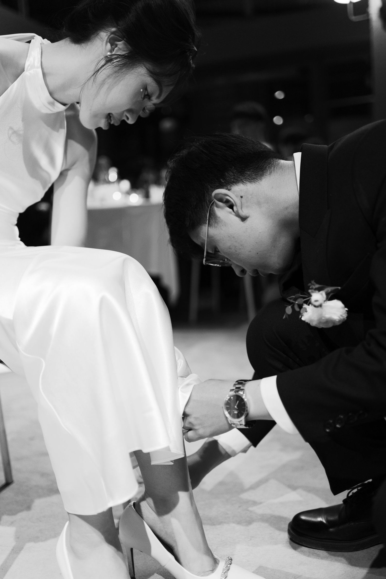 Groom kneeling to place wedding shoe on seated bride in black and white reception photo.