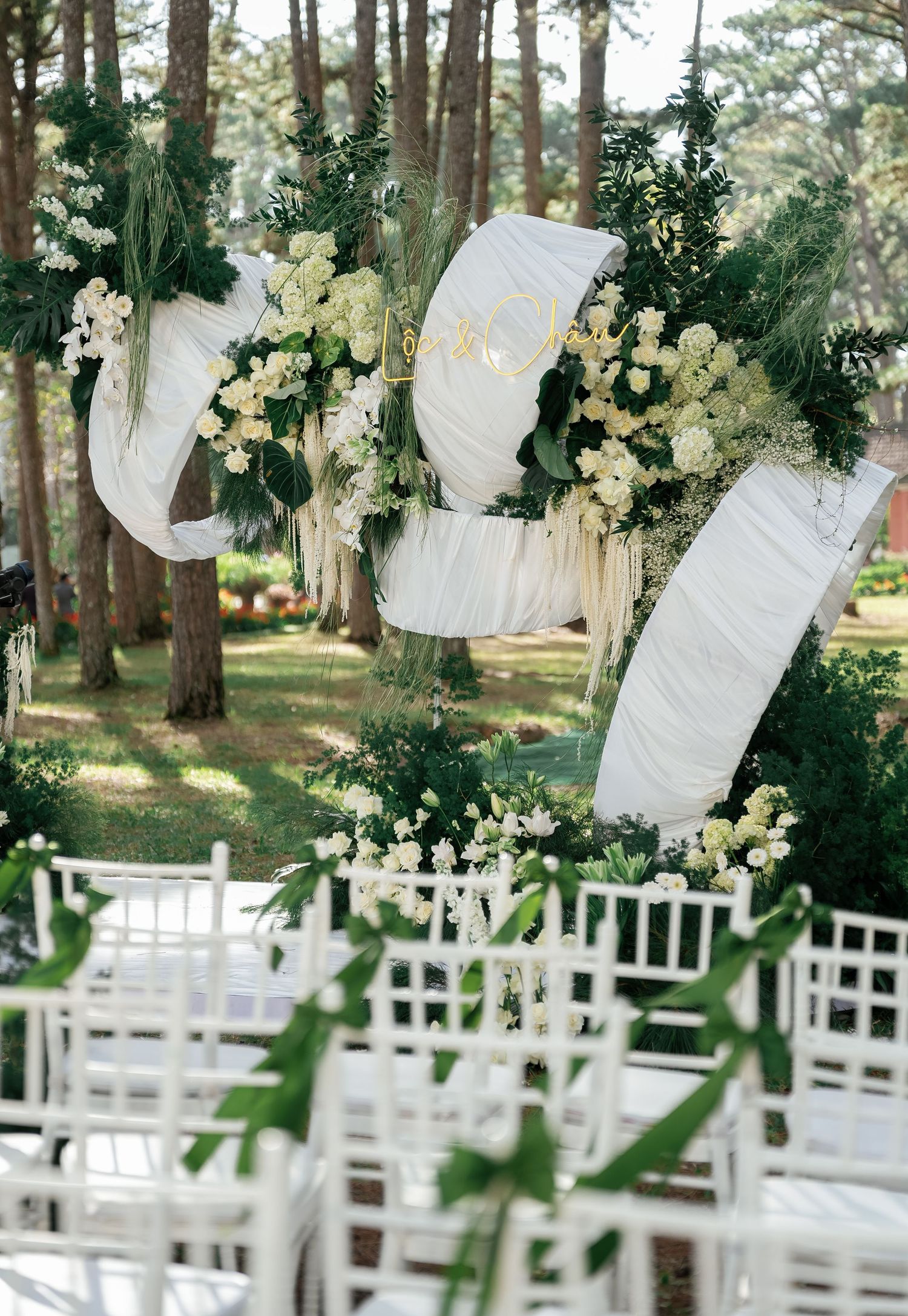 Elegant outdoor wedding ceremony with white draping, lush greenery, and ivory floral arrangements adorning pine trees above white chairs.