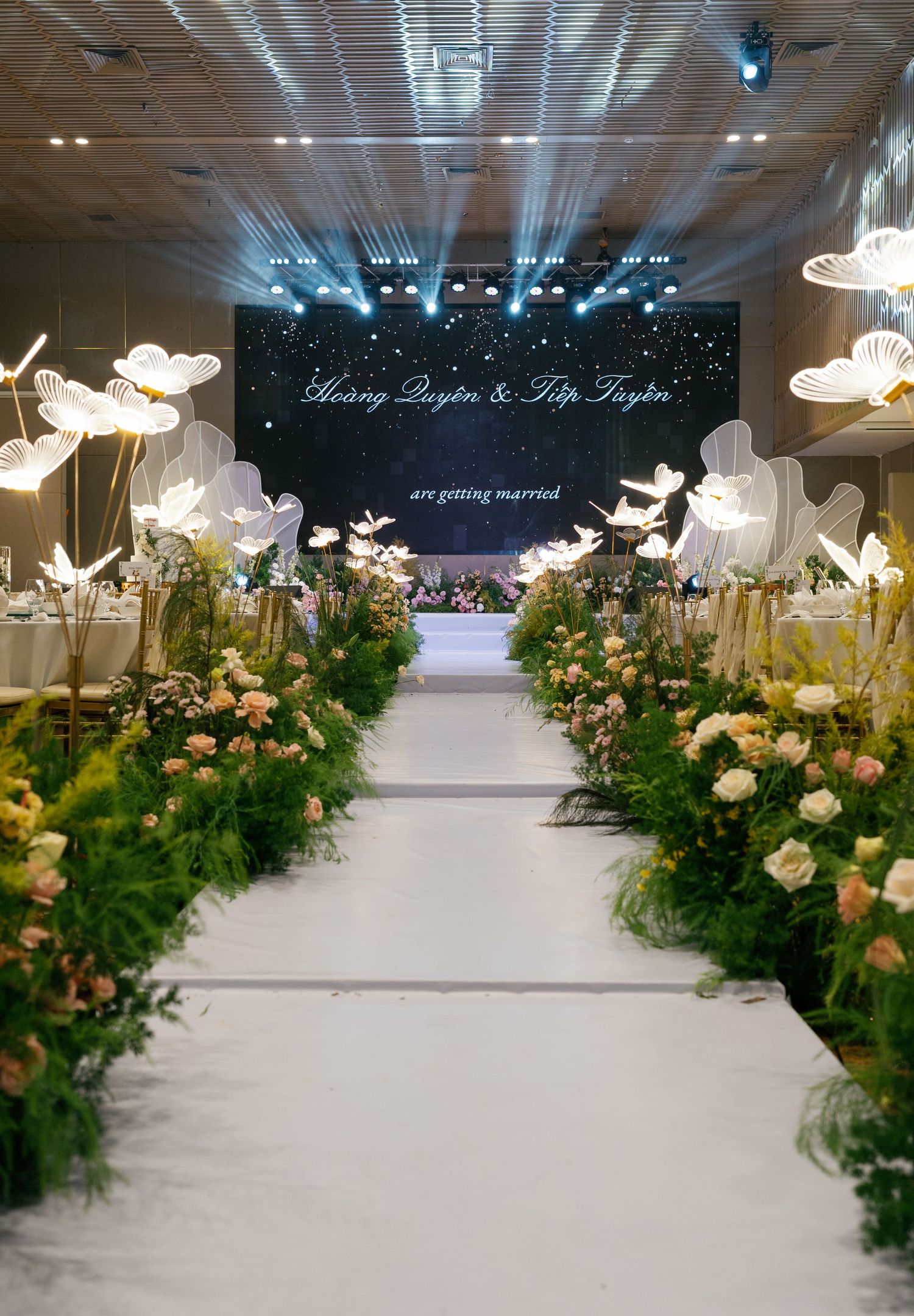 Elegant wedding venue aisle lined with lush greenery, pink roses, and glowing butterfly decorations leading to a dark floral backdrop.