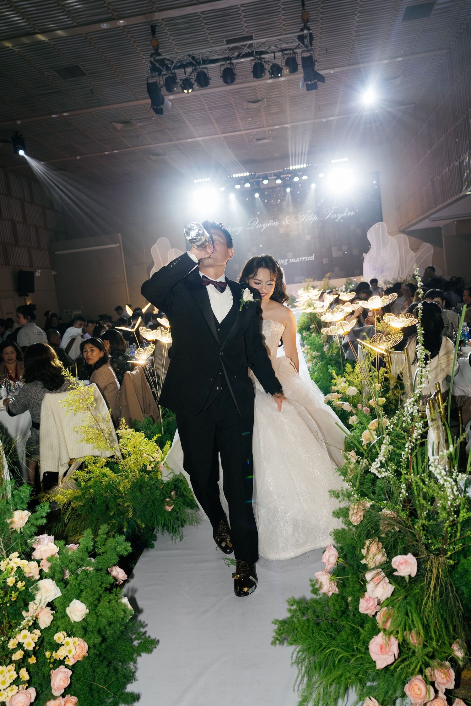 Bride and groom walk down a floral-lined wedding aisle surrounded by glowing butterfly lights and cheering guests.