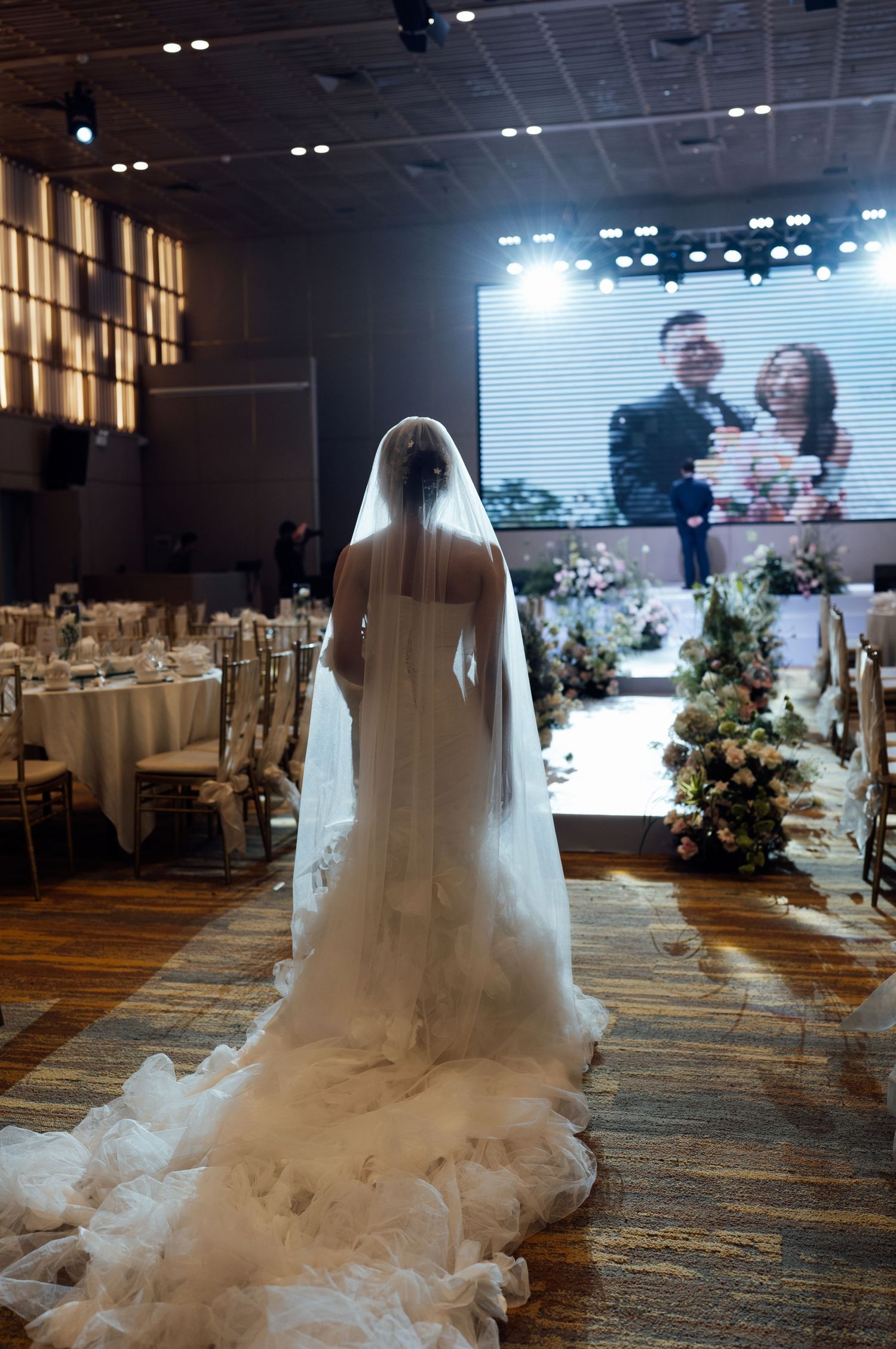 Bride in white gown and veil walks down aisle toward large screen displaying couple at wedding reception.