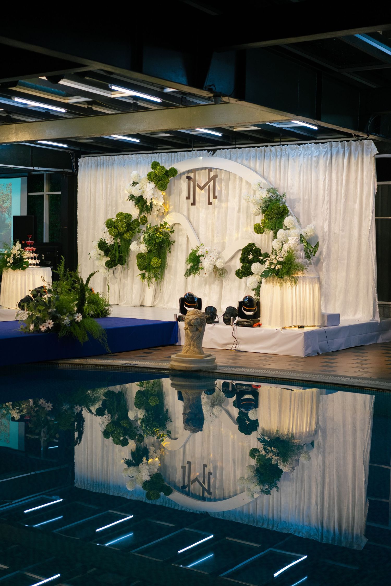Elegant wedding backdrop with monogram, white drapes, green floral decor, reflected in a pool at night.
