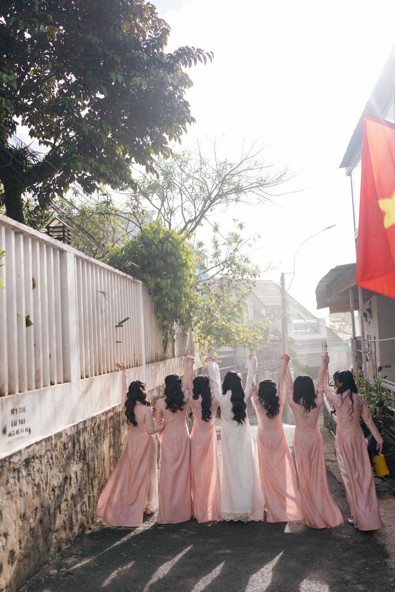 Bridesmaids in pink ao dai gowns raise hands with bride in white dress outdoors near Vietnamese flag.