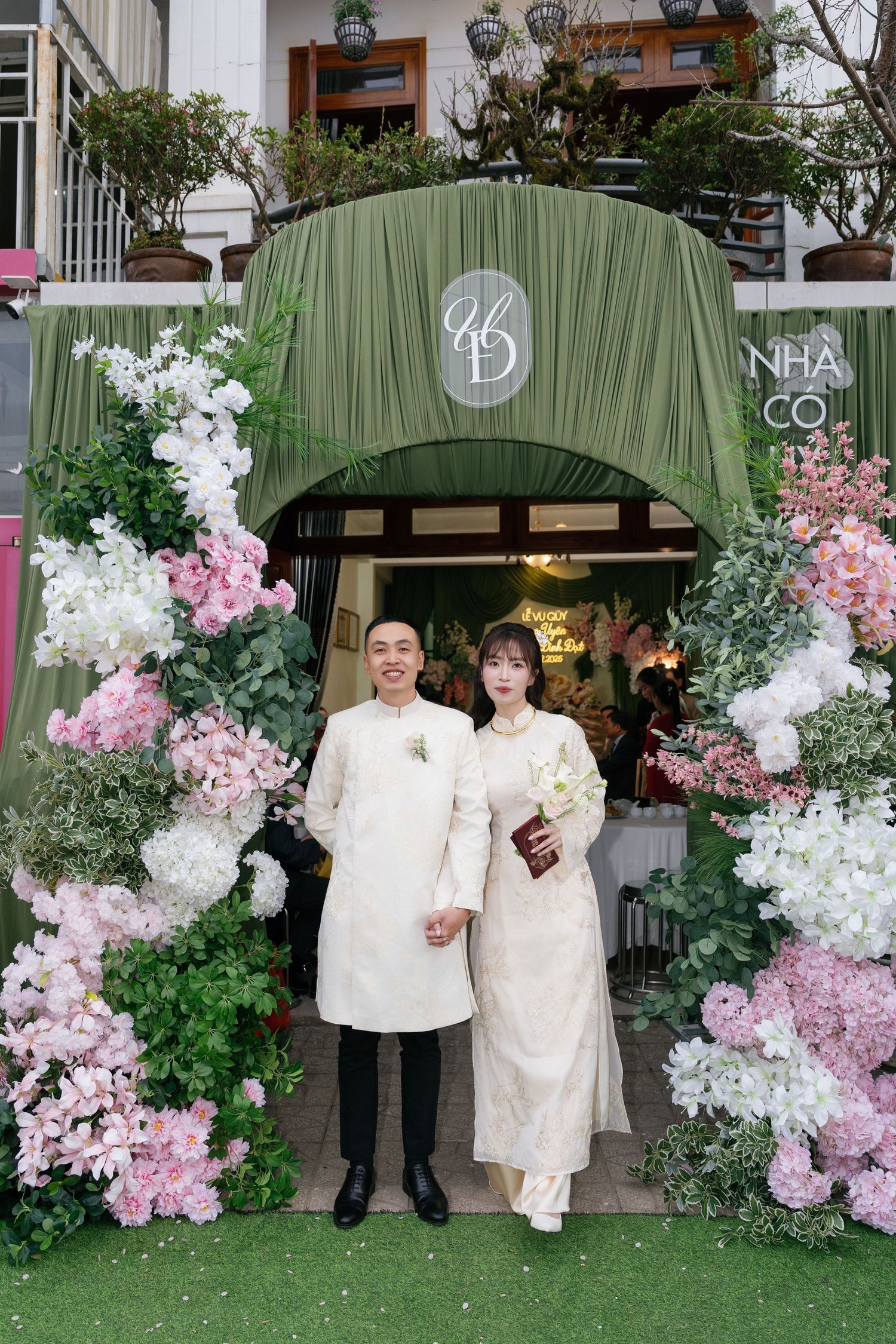 Couple in white traditional Vietnamese ao dai attire posing under floral arch with pink and white blooms at wedding venue.