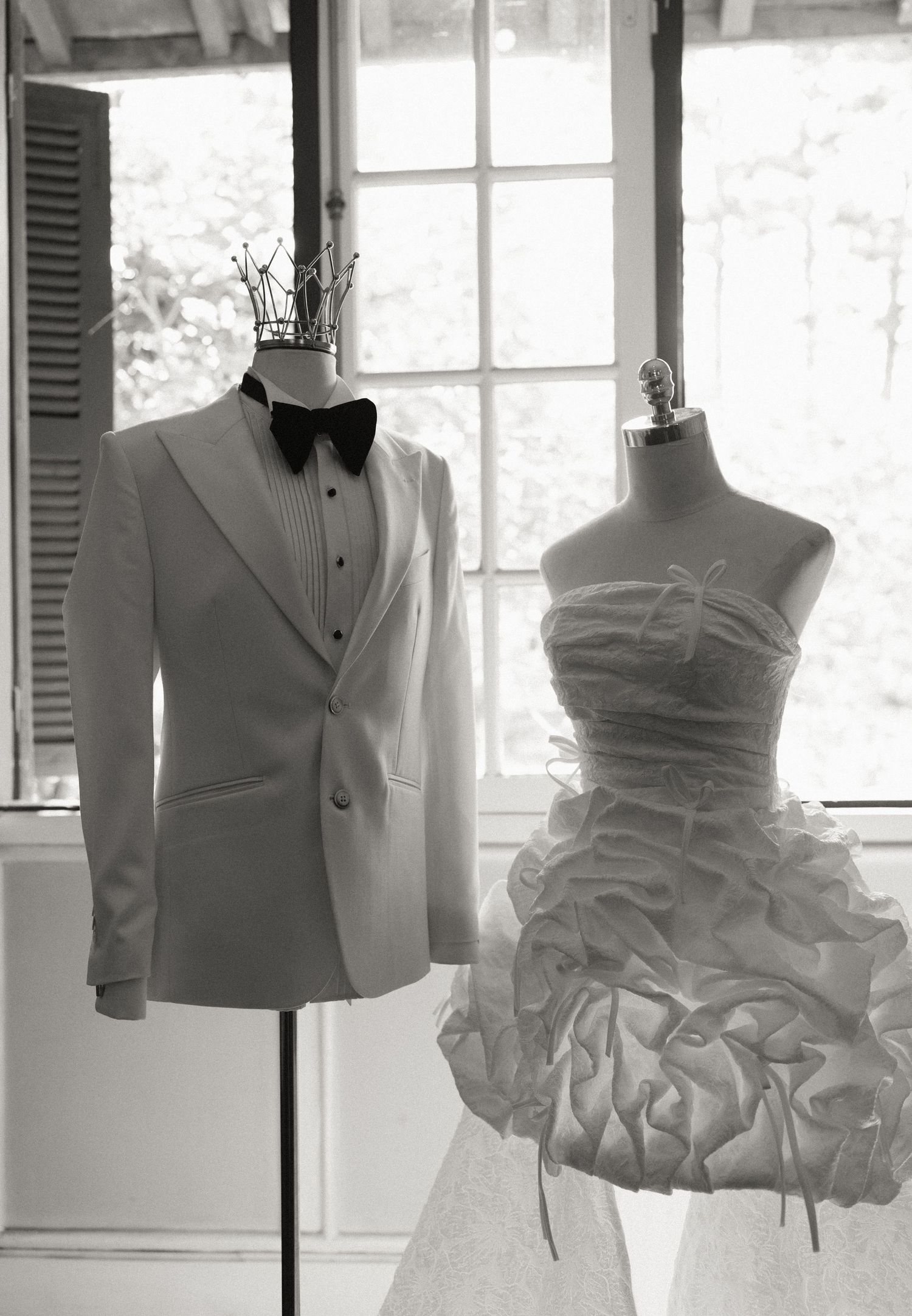 Black and white photo of wedding attire on mannequins, featuring a white tuxedo and a ruffled strapless bridal gown.