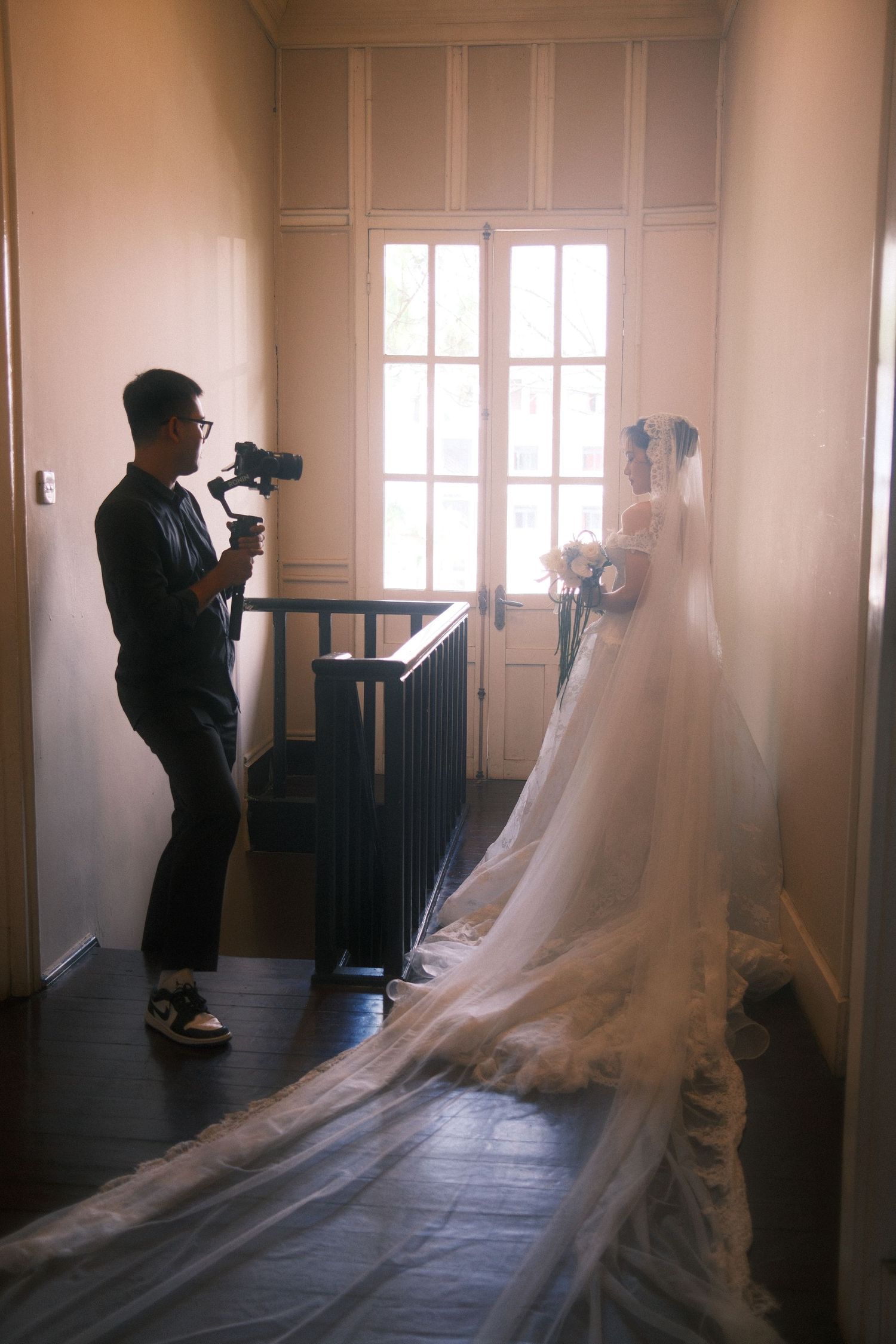 Videographer filming a bride in a white wedding gown with long veil near a window in an elegant hallway.