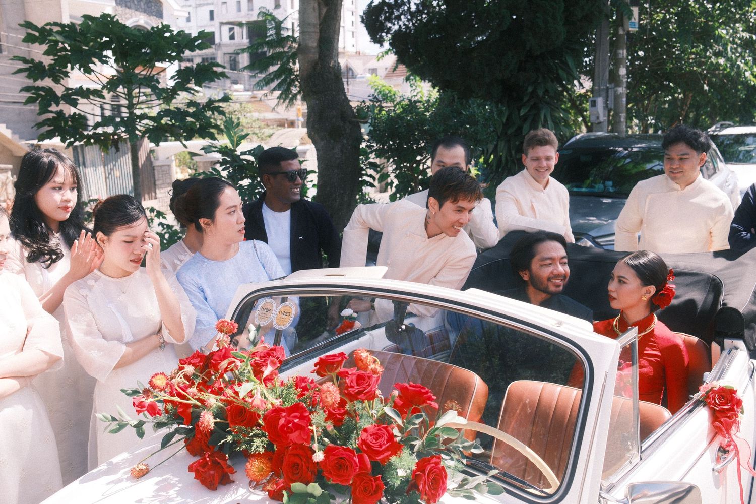 A joyful wedding party gathers around a classic white convertible adorned with red roses, celebrating outdoors.