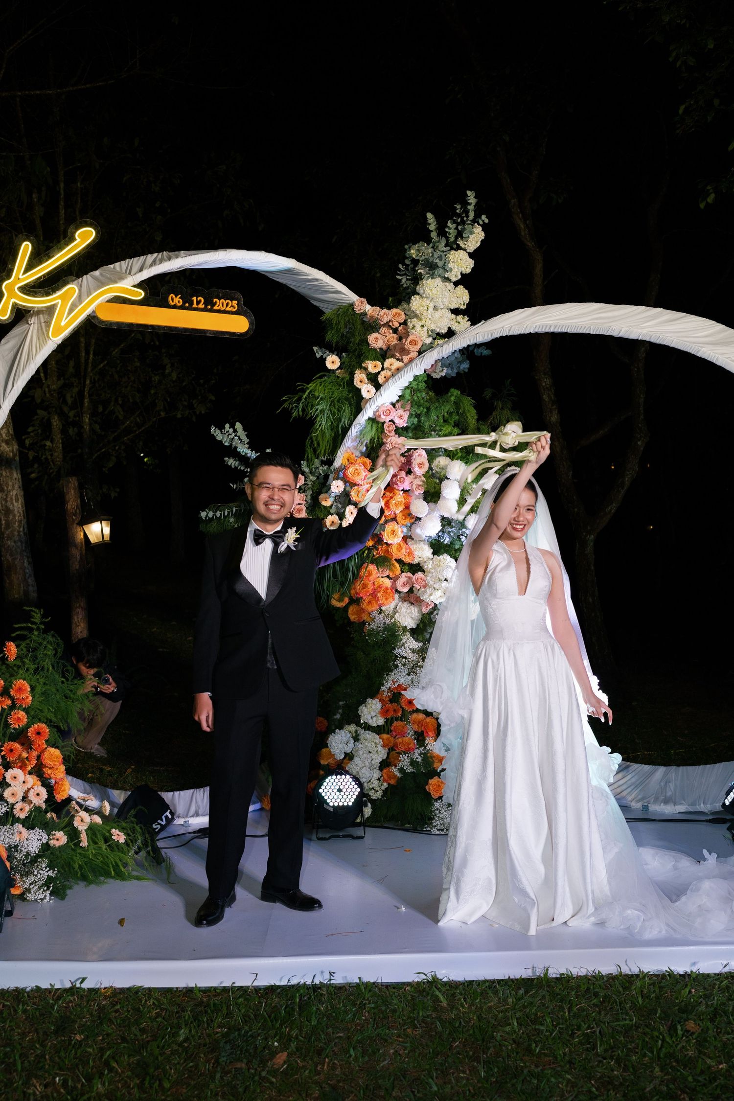 Smiling bride and groom pose under white curved arch adorned with flowers at night ceremony.
