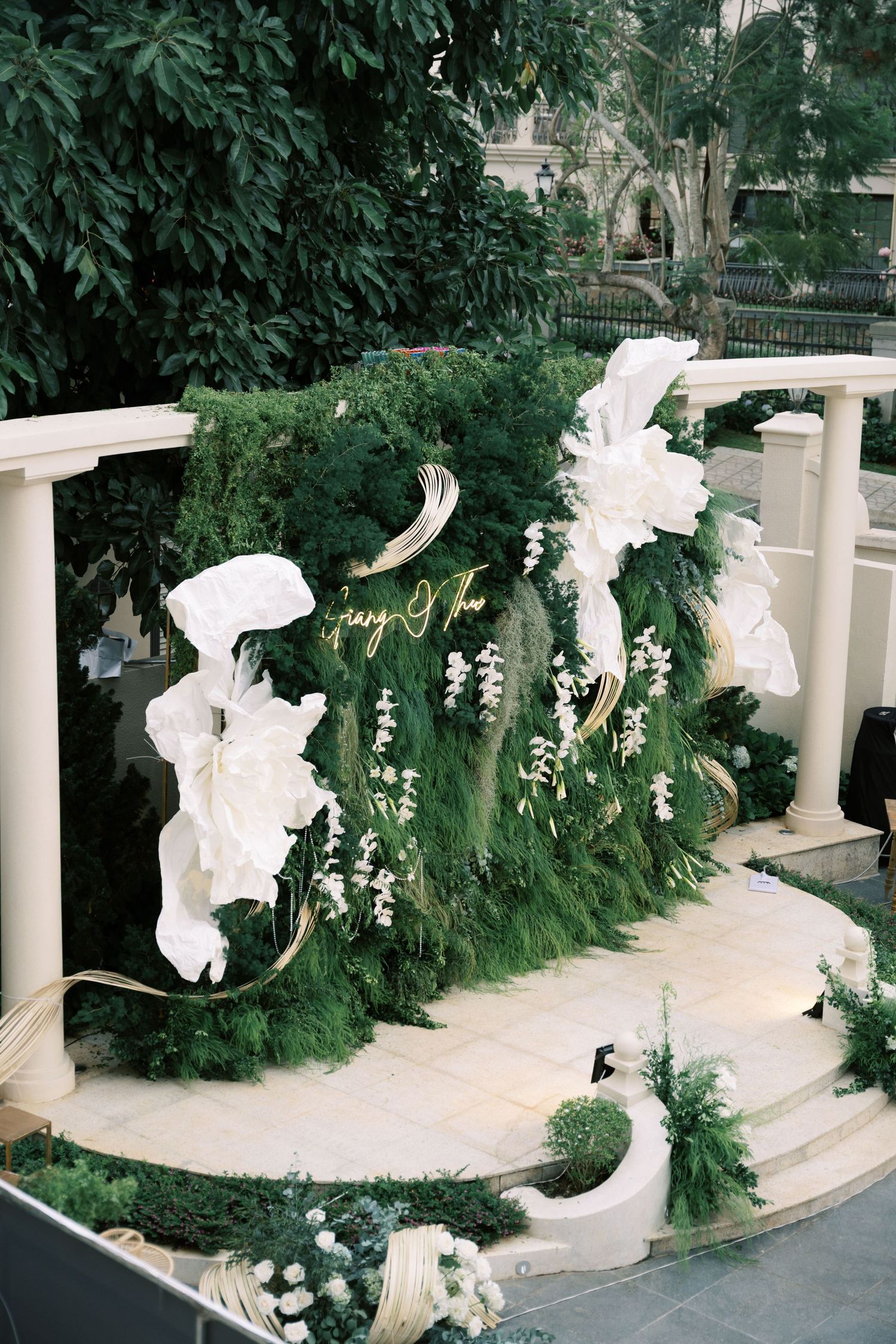 Lush green wedding backdrop adorned with oversized white paper flowers and gold accents outdoors.