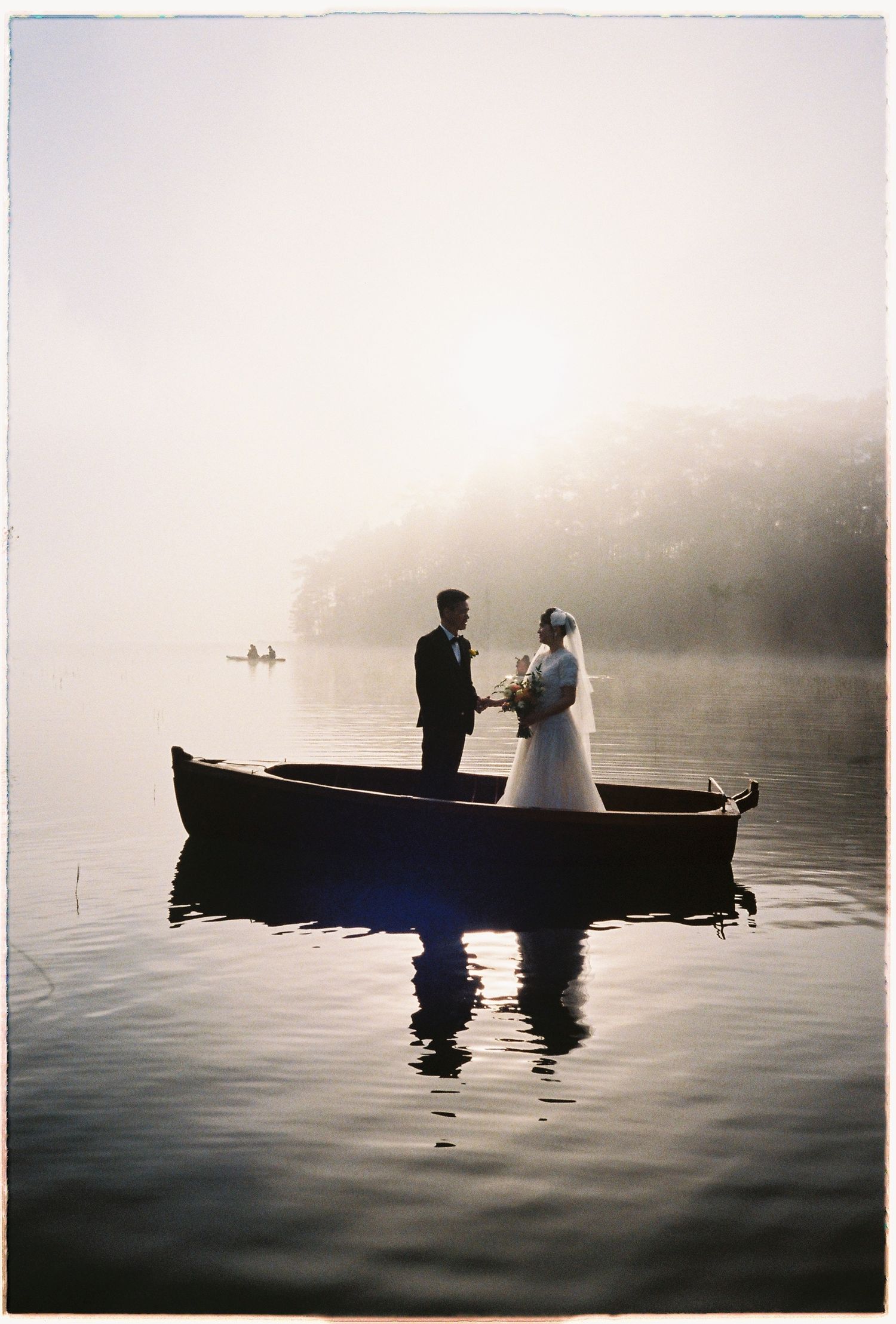 A bride and groom hold hands in a wooden boat on a serene, fog-covered lake at dawn.