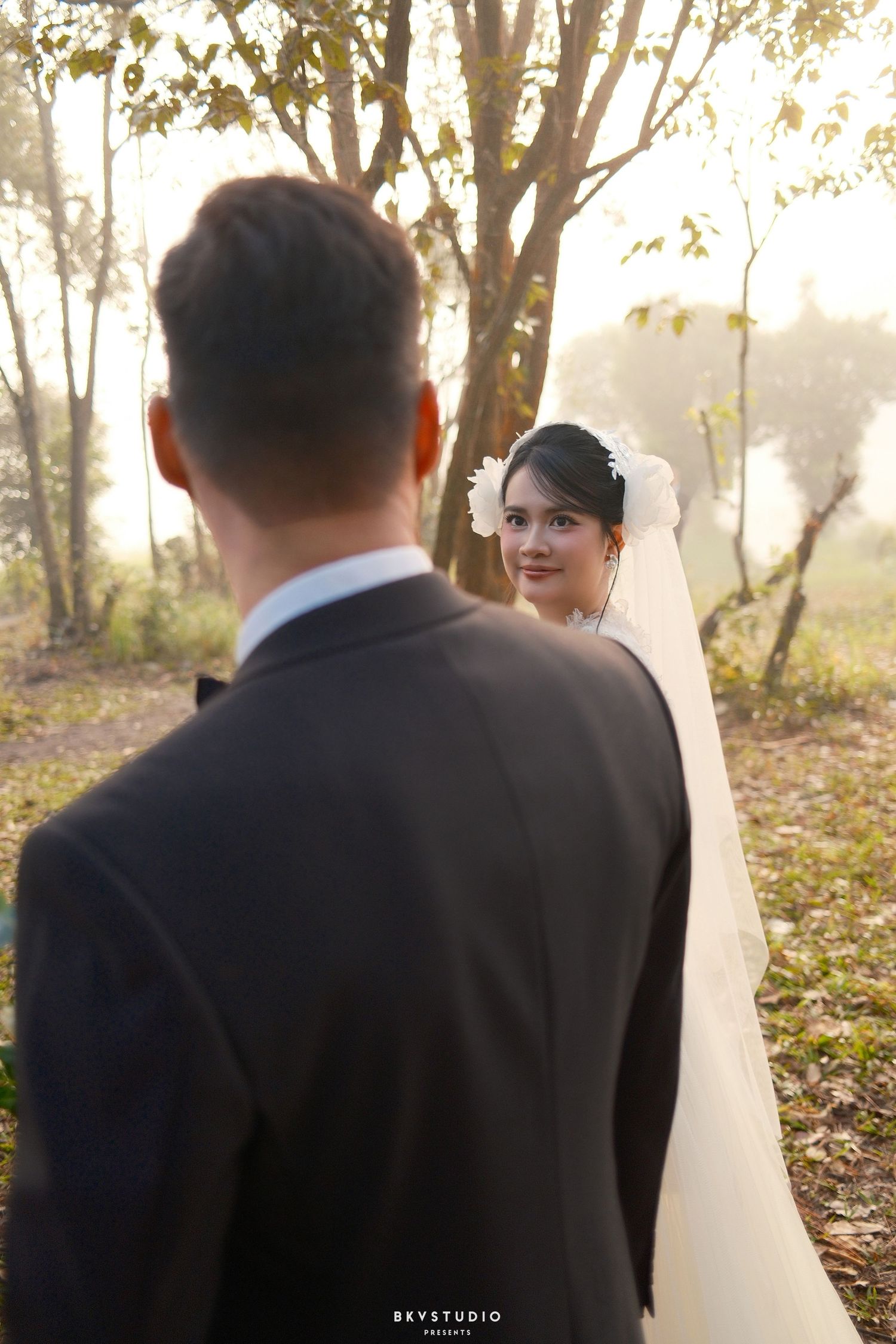 A groom in a dark suit faces a smiling bride in a white veil among sunlit trees.