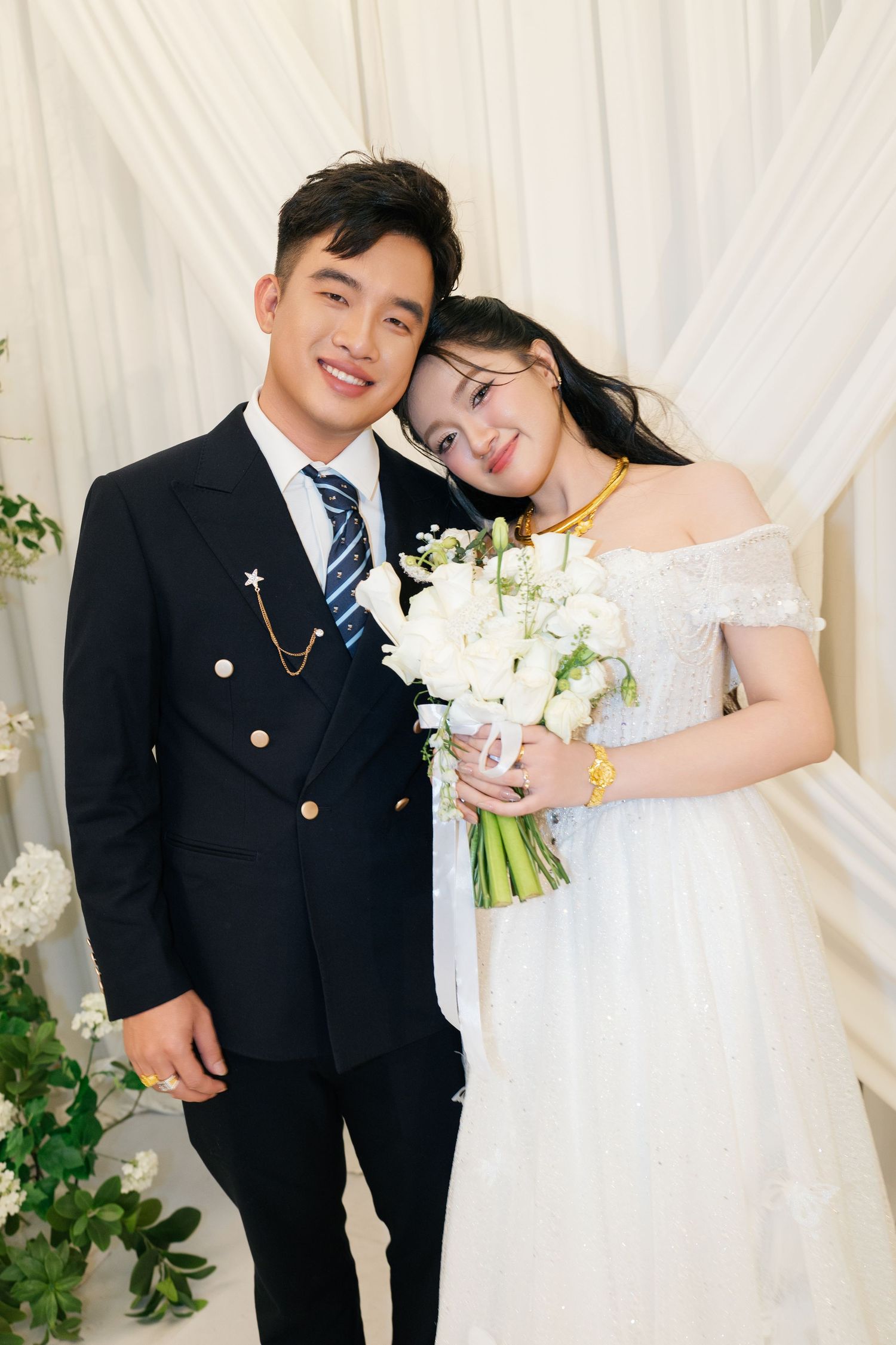 Vietnamese couple in wedding attire pose together, groom in black suit, bride in white off-shoulder gown.