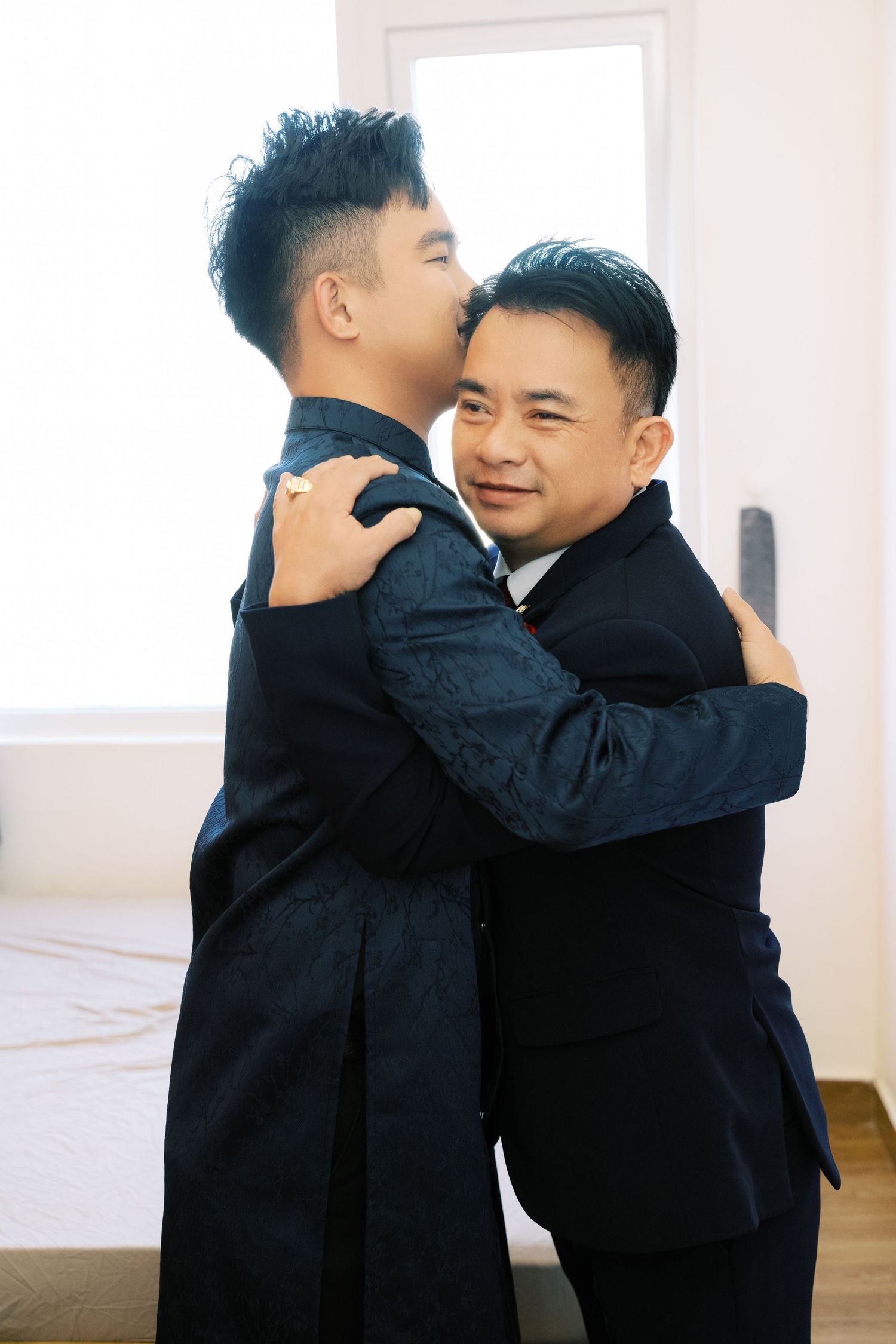 Two Vietnamese men in dark suits sharing an embrace at a wedding celebration.