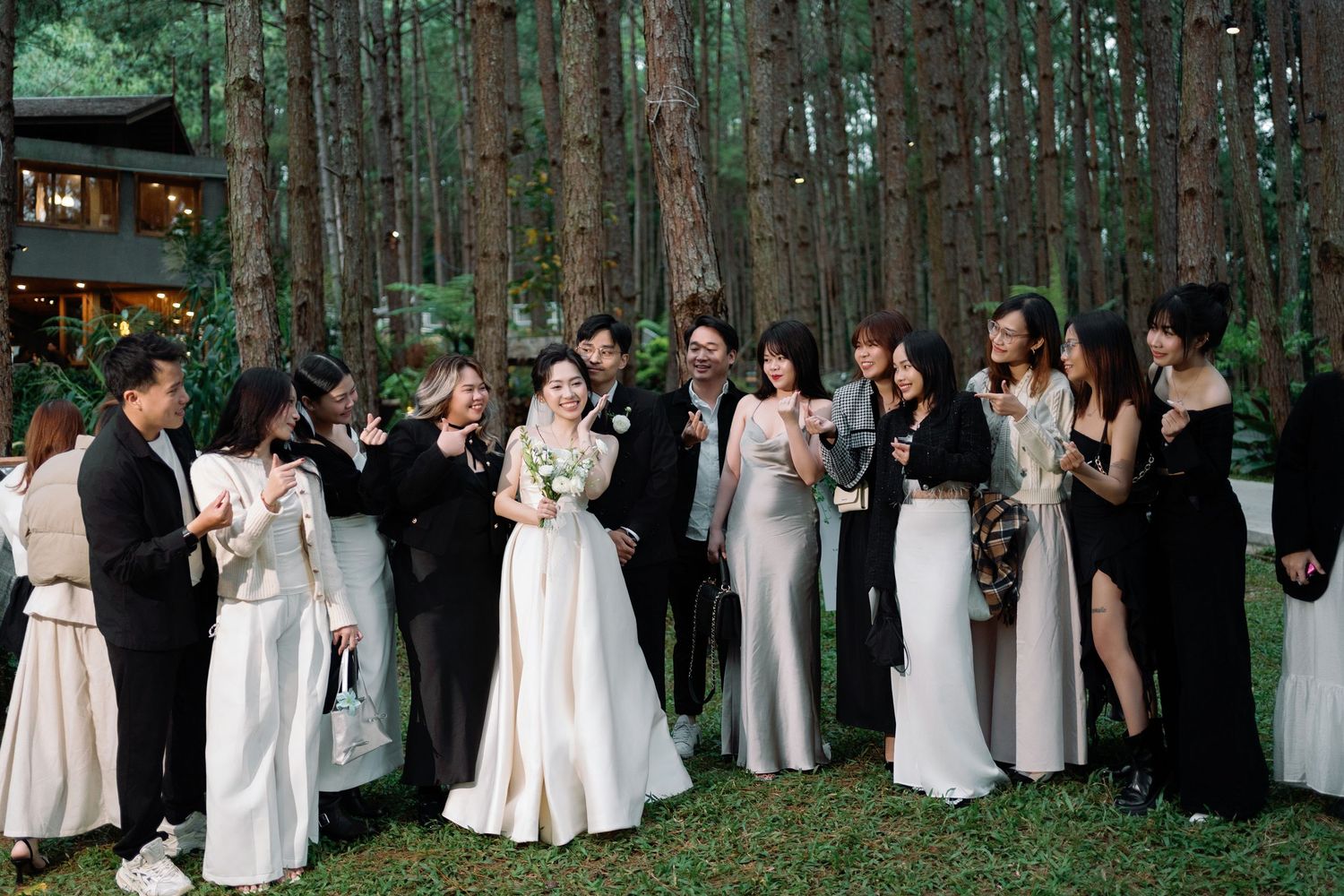 Wedding party celebrating outdoors in pine forest with bride in white gown and guests in black attire.