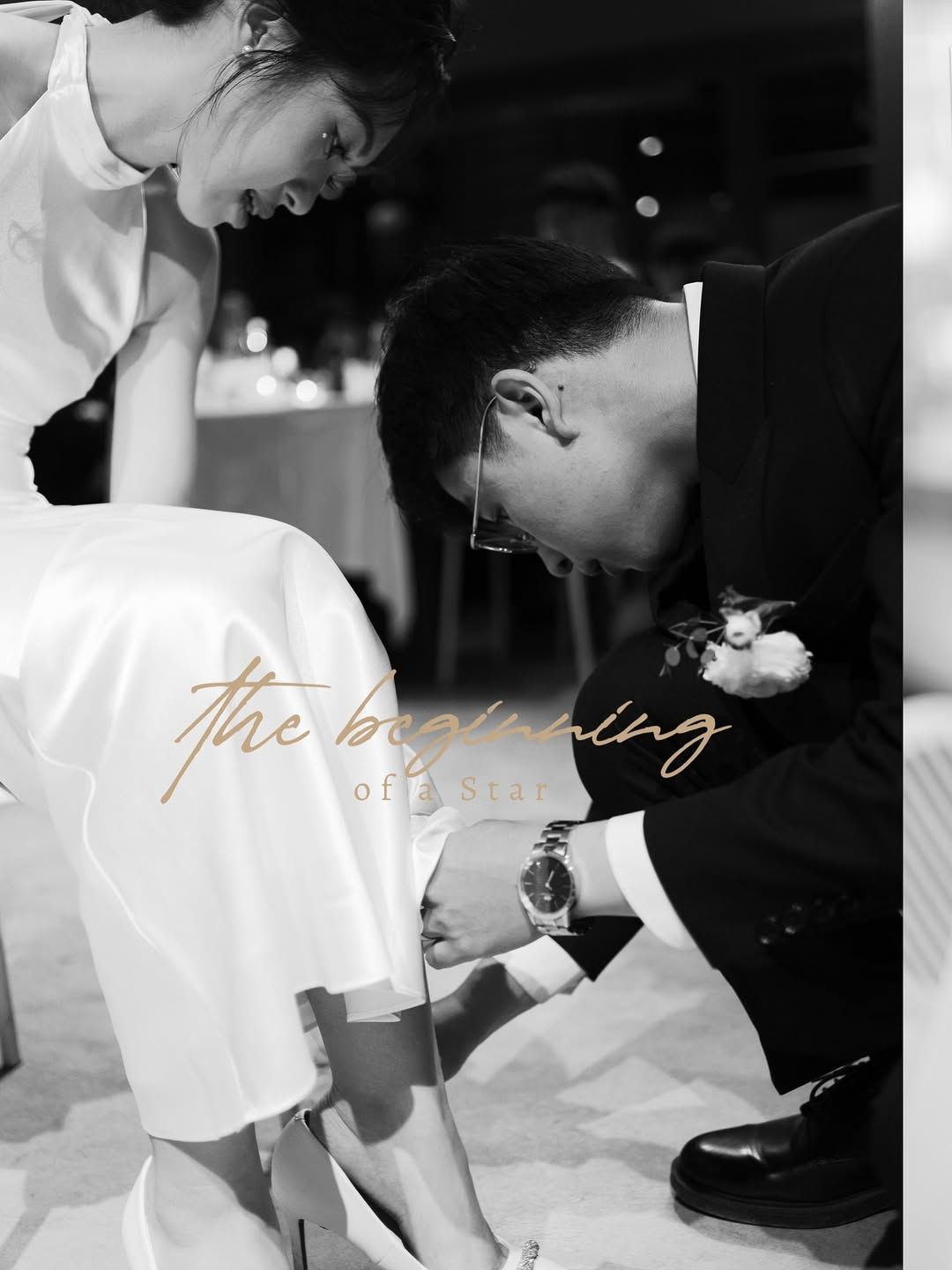 Groom kneeling to place shoe on bride's foot at wedding reception, black and white photo.