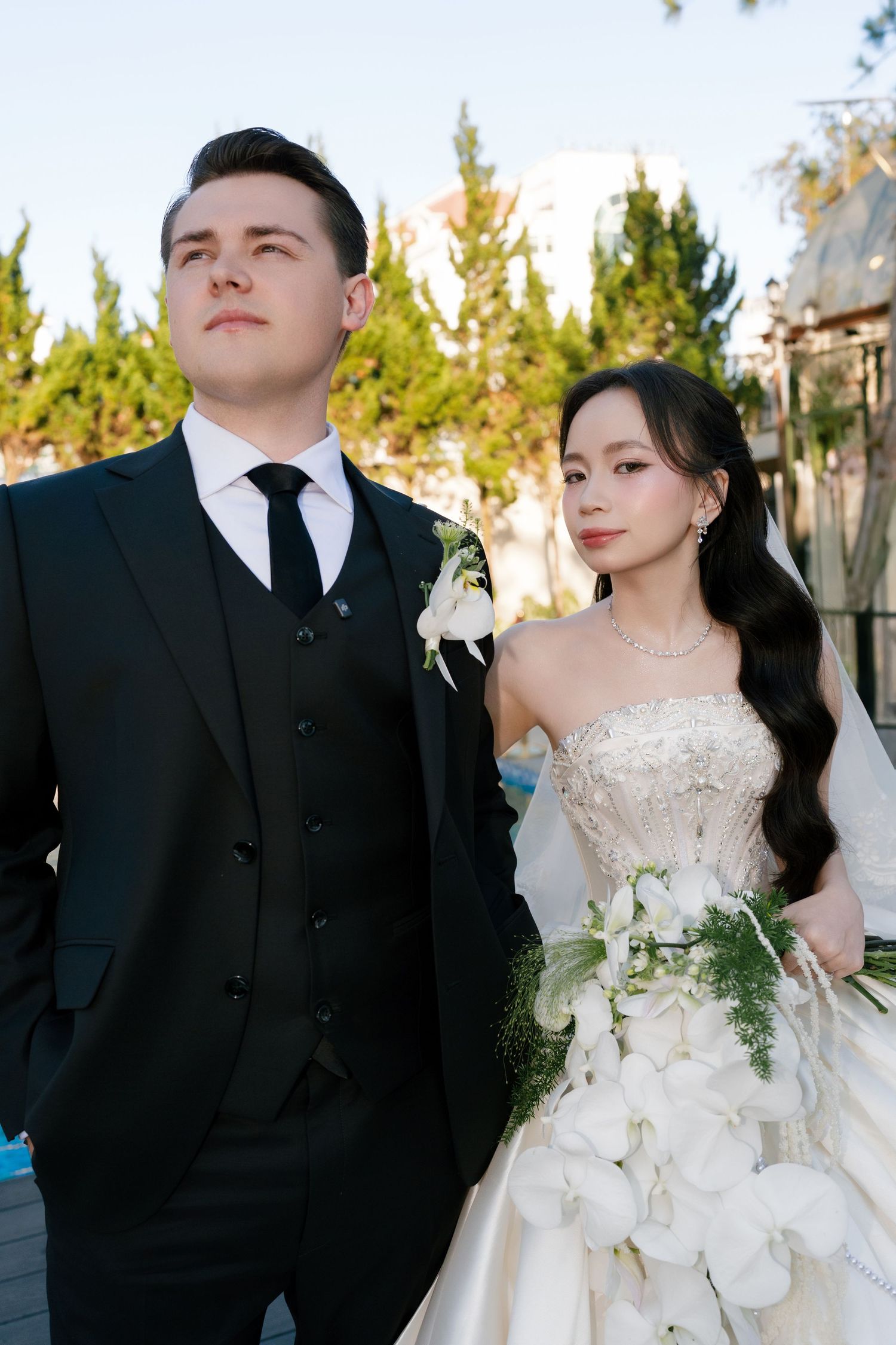 Elegant wedding couple posing outdoors, groom in black suit, bride in white strapless gown holding white floral bouquet.