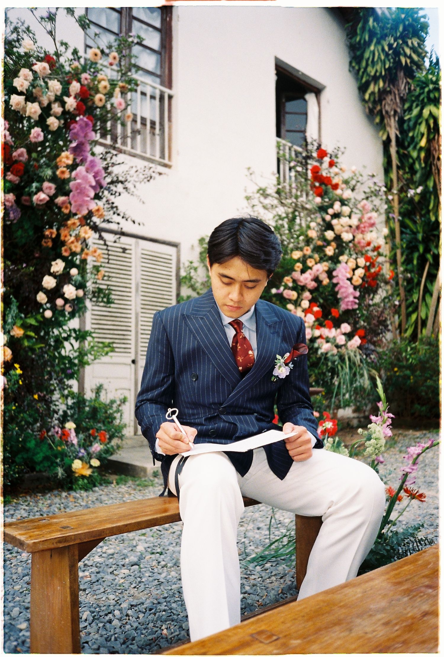 A man in a navy pinstripe suit reads papers while seated on a bench surrounded by colorful garden flowers.