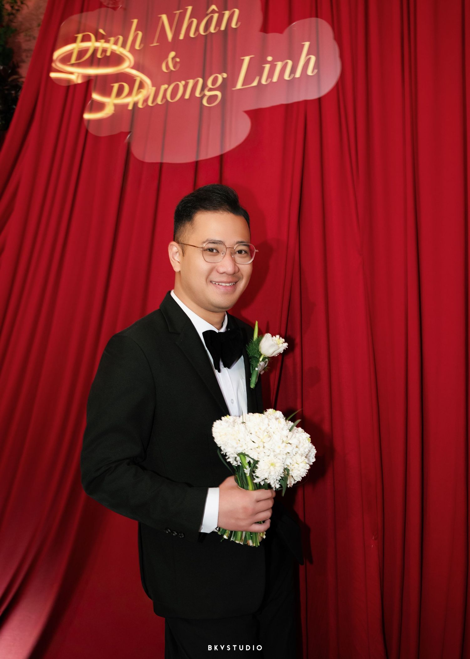 Groom in black tuxedo holding white flower bouquet at Dinh Nhan and Phuong Linh wedding ceremony.
