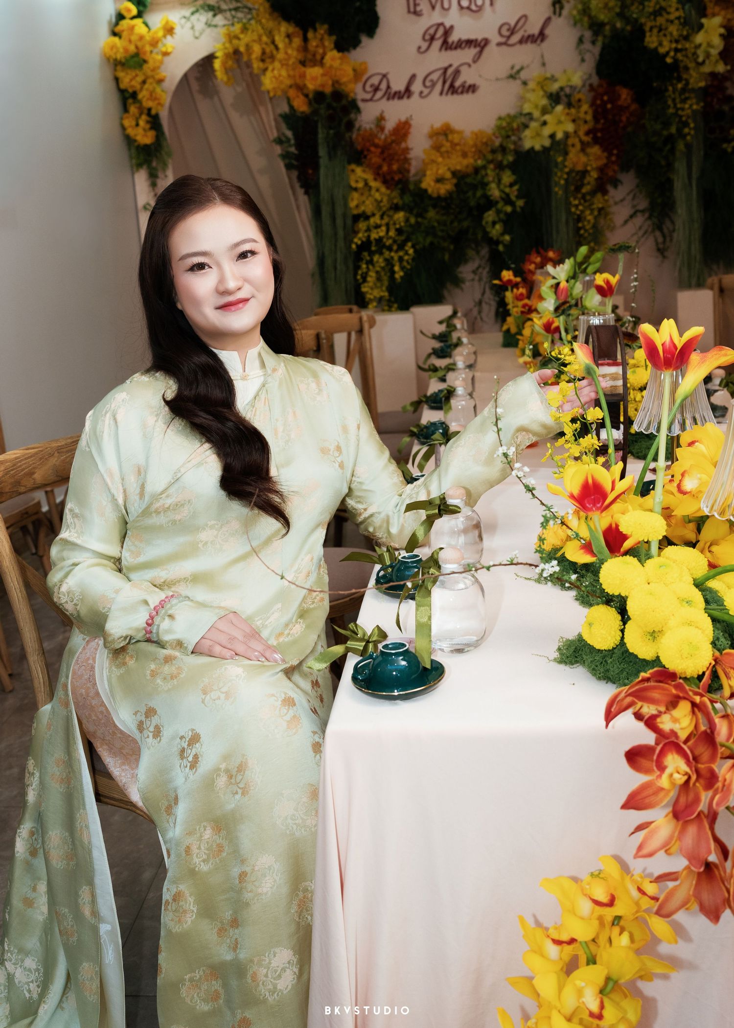 A woman in a light green floral ao dai sits gracefully at a table adorned with vibrant yellow and red flower arrangements.