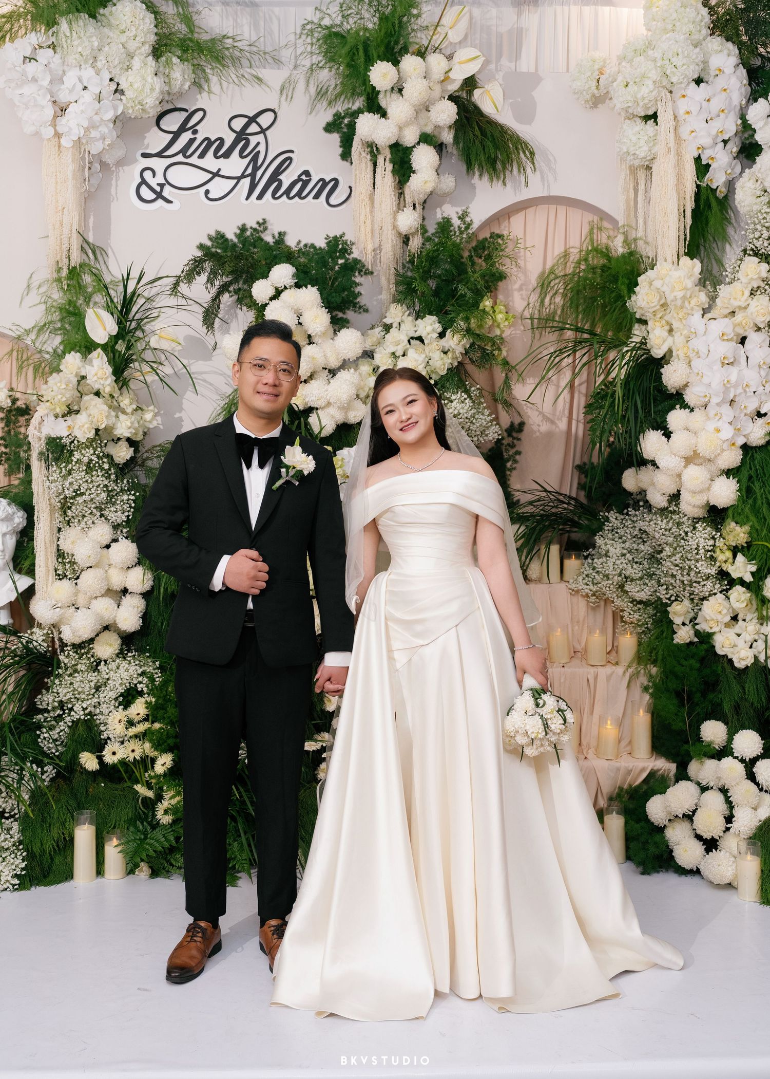Couple poses at elegant wedding backdrop with white floral arrangements, greenery, and a sign reading Linh & Nhan.