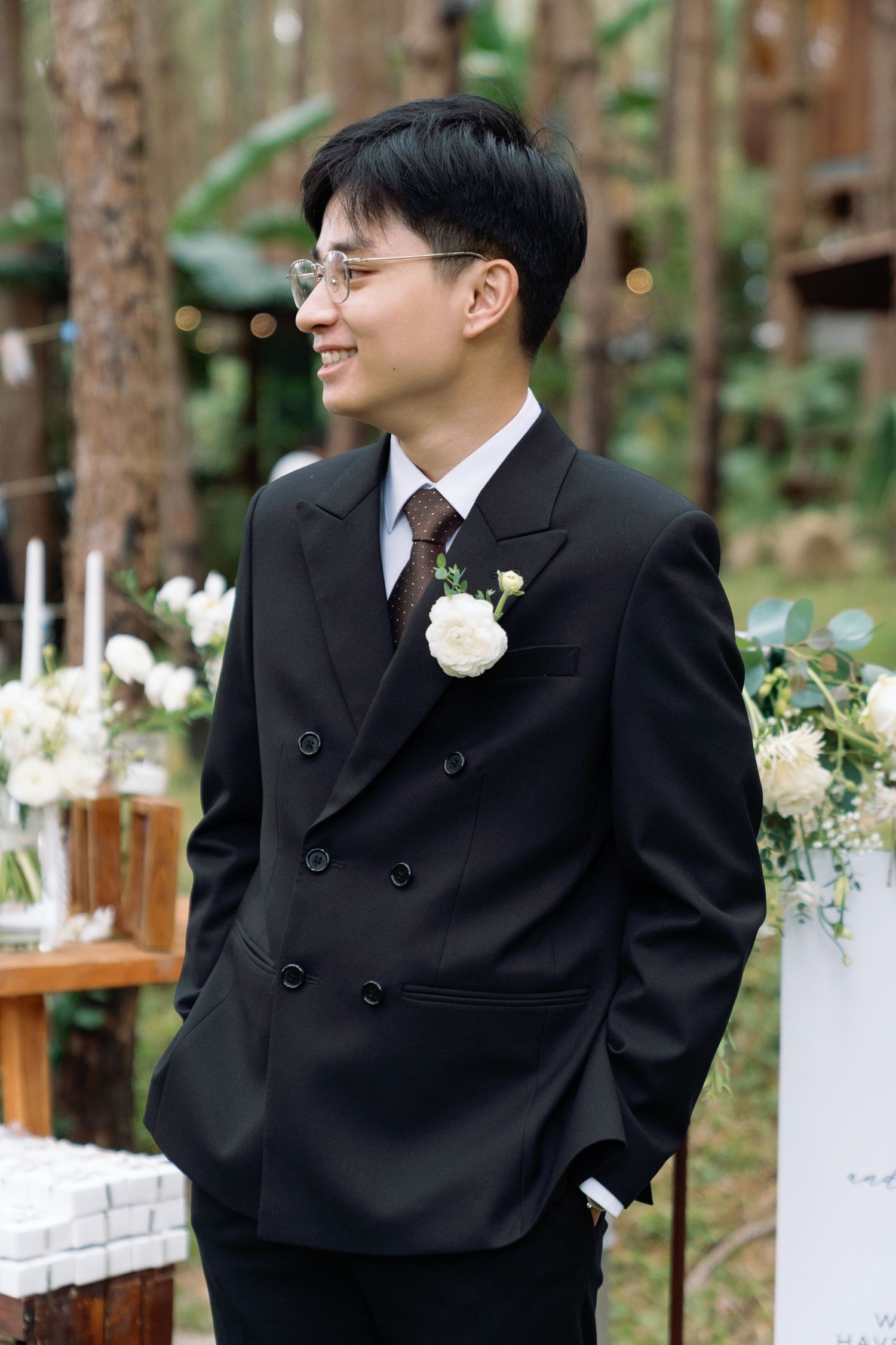 Smiling groom in black double-breasted suit with white boutonniere standing at outdoor forest wedding ceremony.