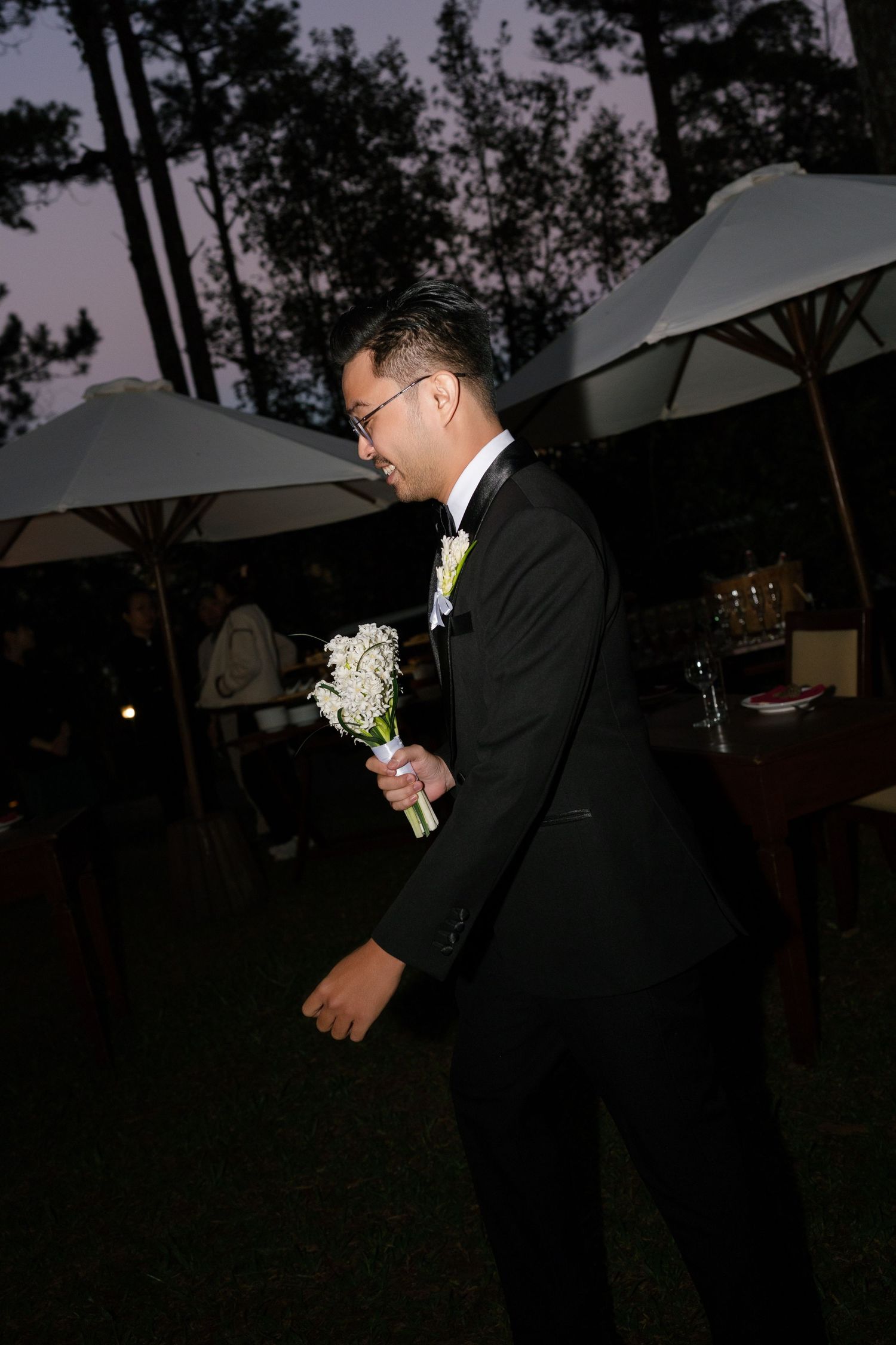 Groom in black tuxedo holding white bridal bouquet outdoors at night with white umbrellas in background.