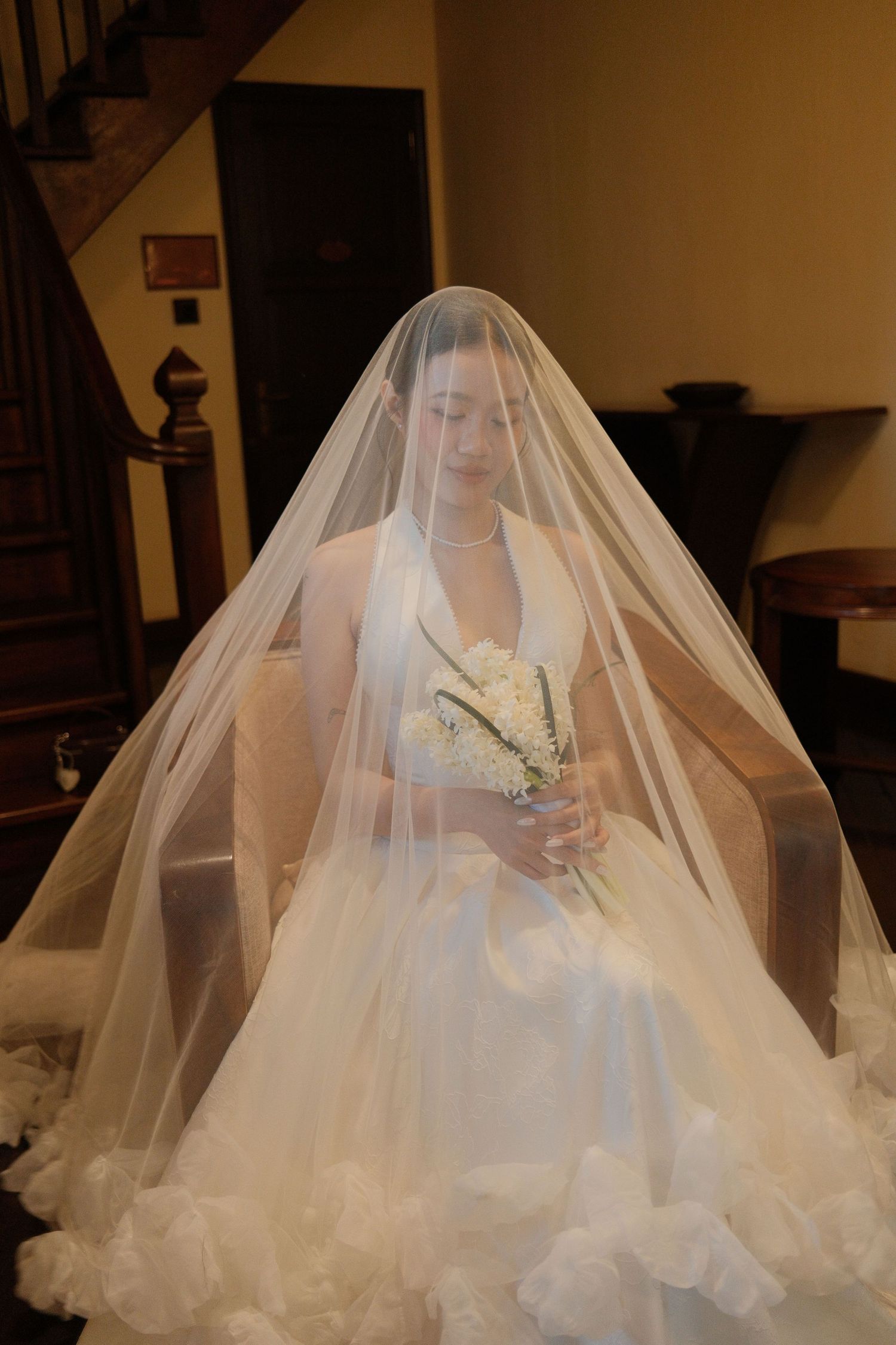 A veiled bride in a white ruffled gown holds a white bouquet while seated in a vintage interior.