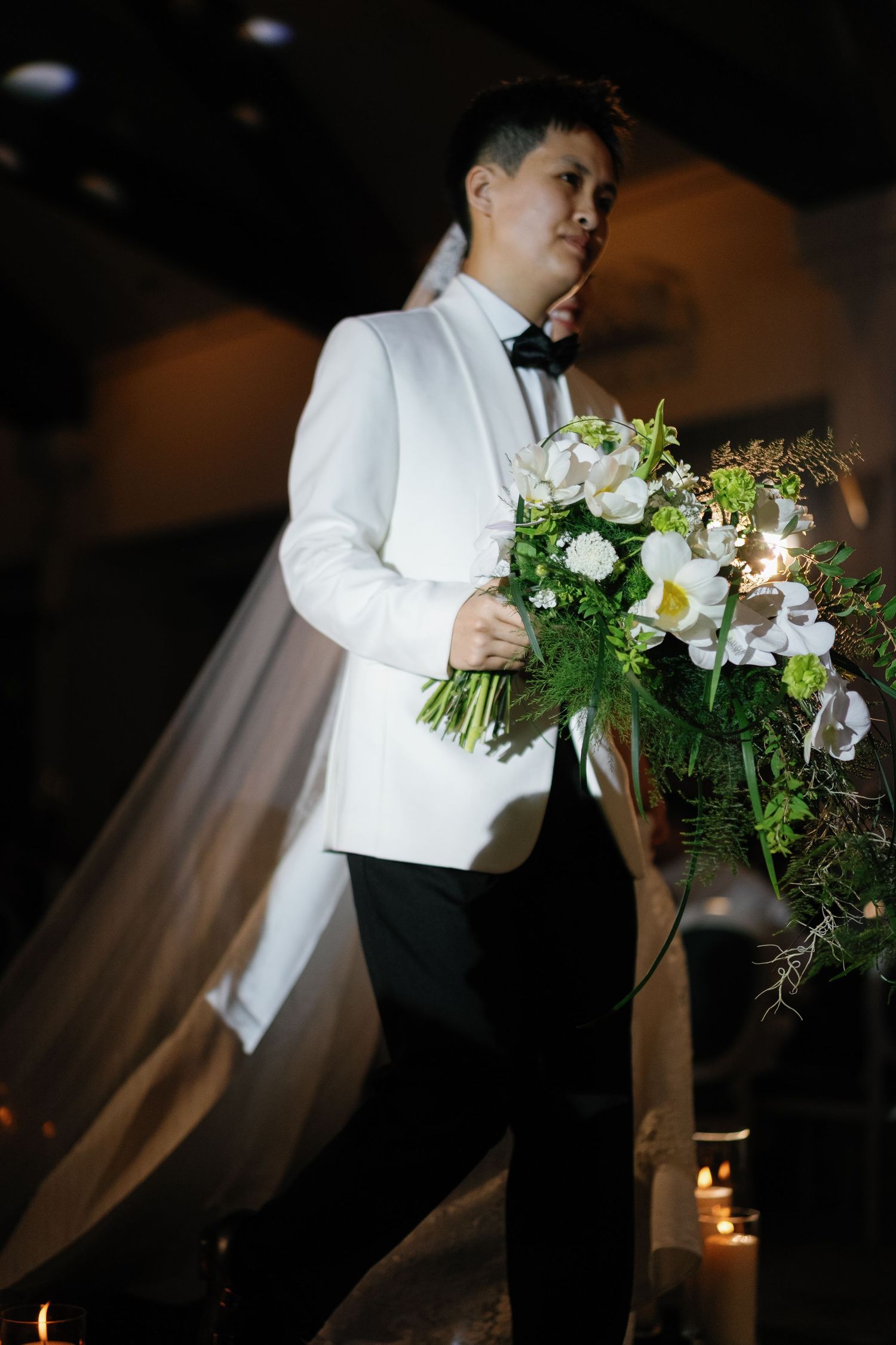 Groom in white tuxedo jacket holding large white floral bouquet at elegant wedding reception.