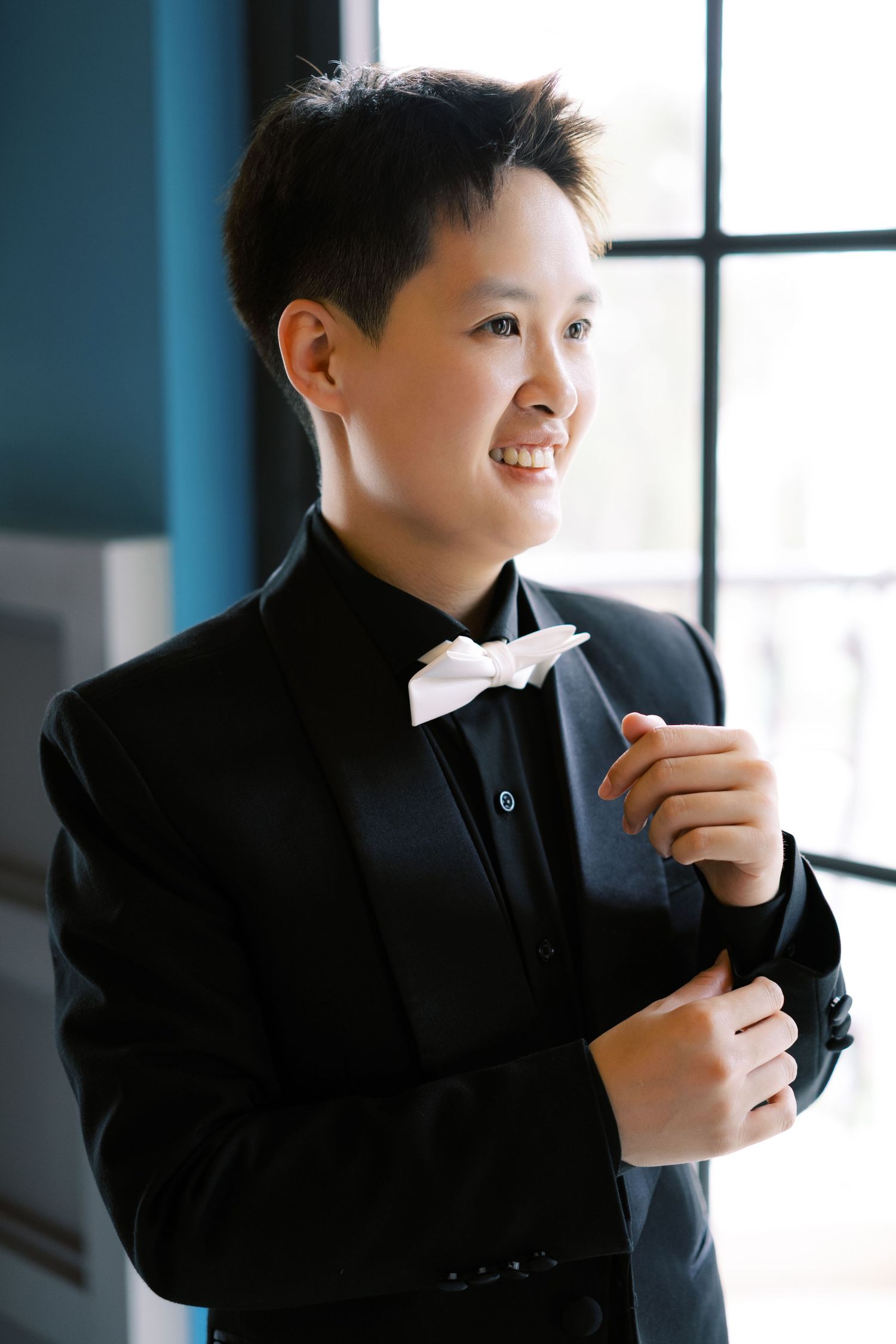 Smiling young Asian person in black tuxedo with white bow tie adjusting jacket near window.