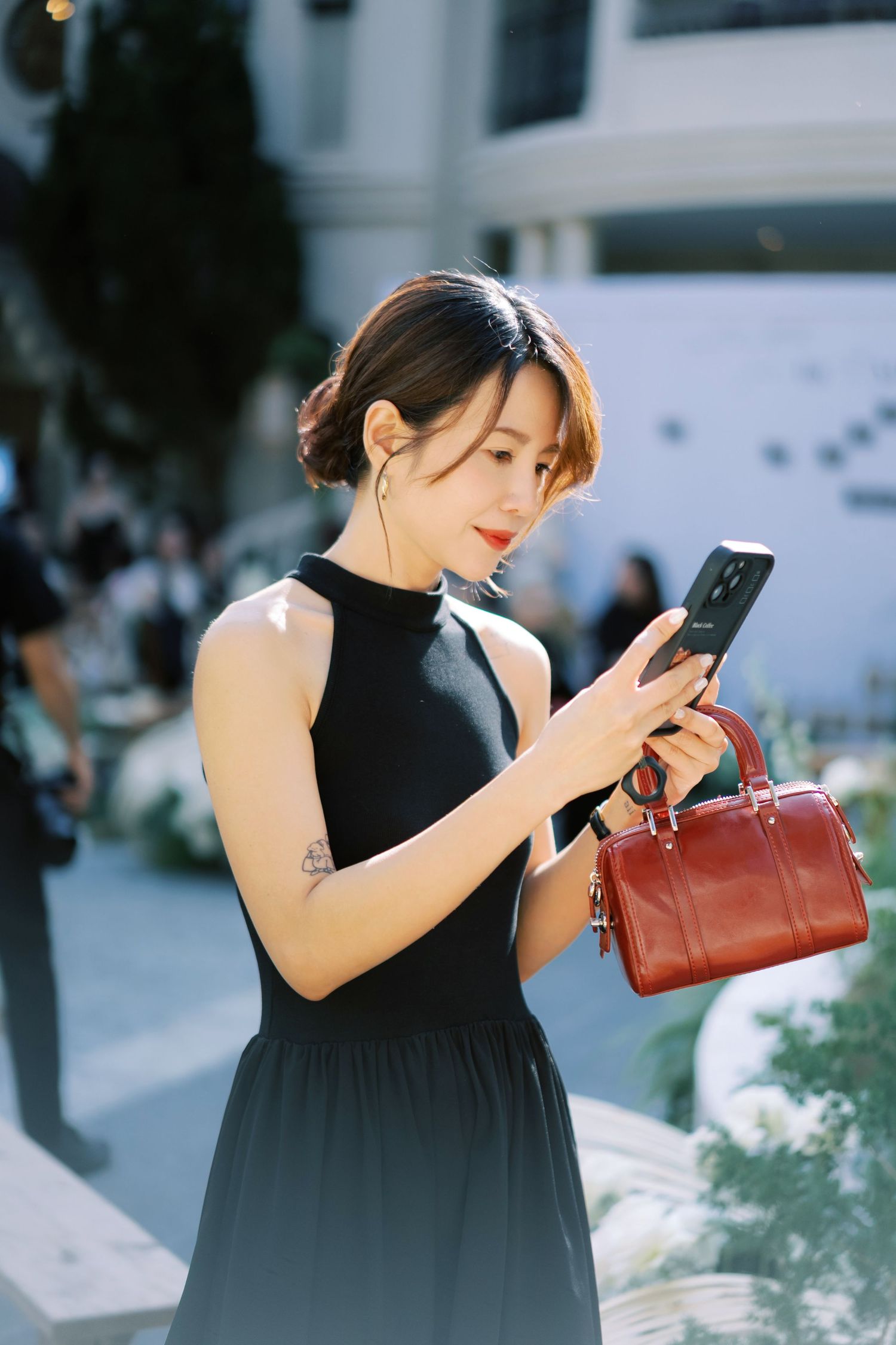 Stylish woman in black sleeveless dress holding a brown handbag while looking at her smartphone outdoors.