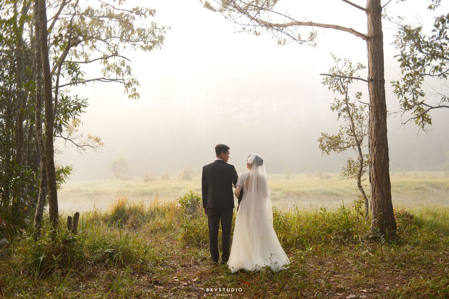 Bride and groom stand hand in hand facing a misty forest landscape on their wedding day.