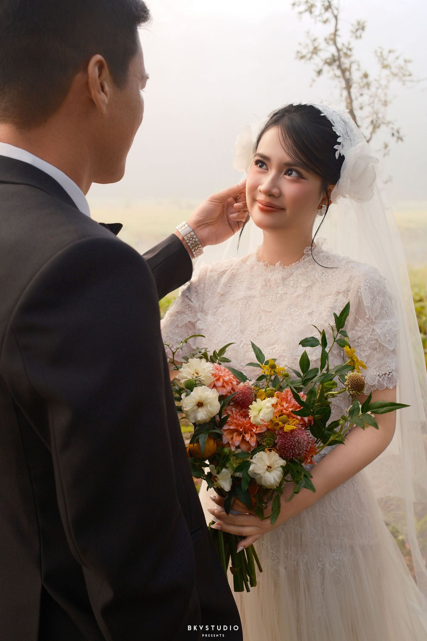 Groom tenderly touches bride's face as she smiles holding a vibrant bouquet in a misty outdoor setting.