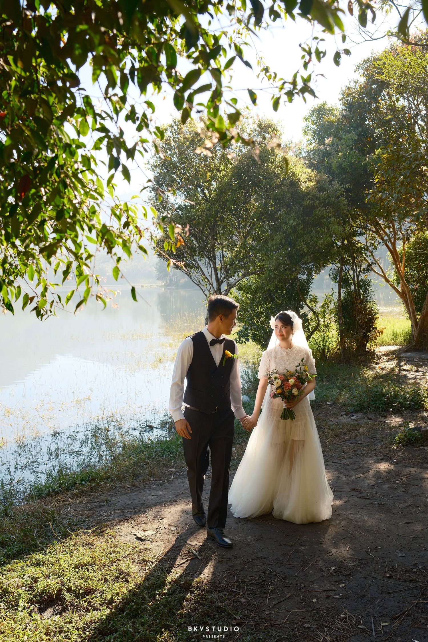 Bride and groom walking hand in hand along a lakeside path surrounded by trees.