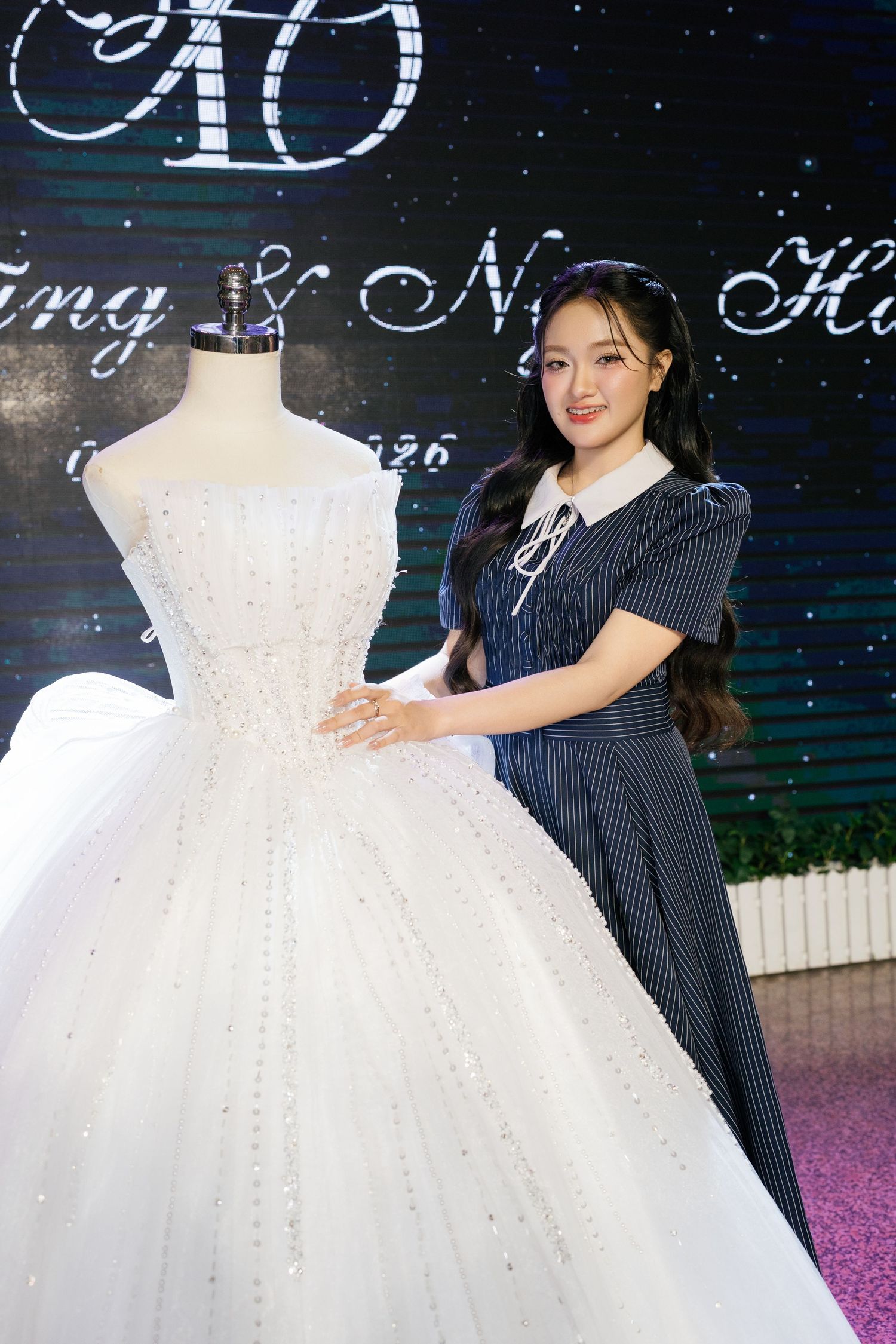 Young Asian woman in striped dress posing beside an elegant white ballgown on a mannequin at a wedding event.